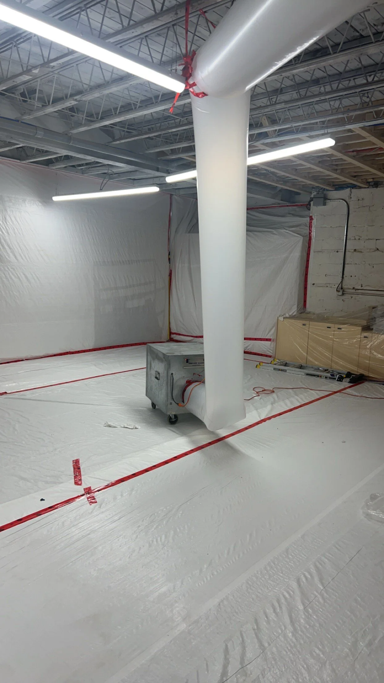 Construction site with white plastic sheeting covering the floor and walls, a large white duct hanging from the ceiling, and a portable heater visible in the center.
