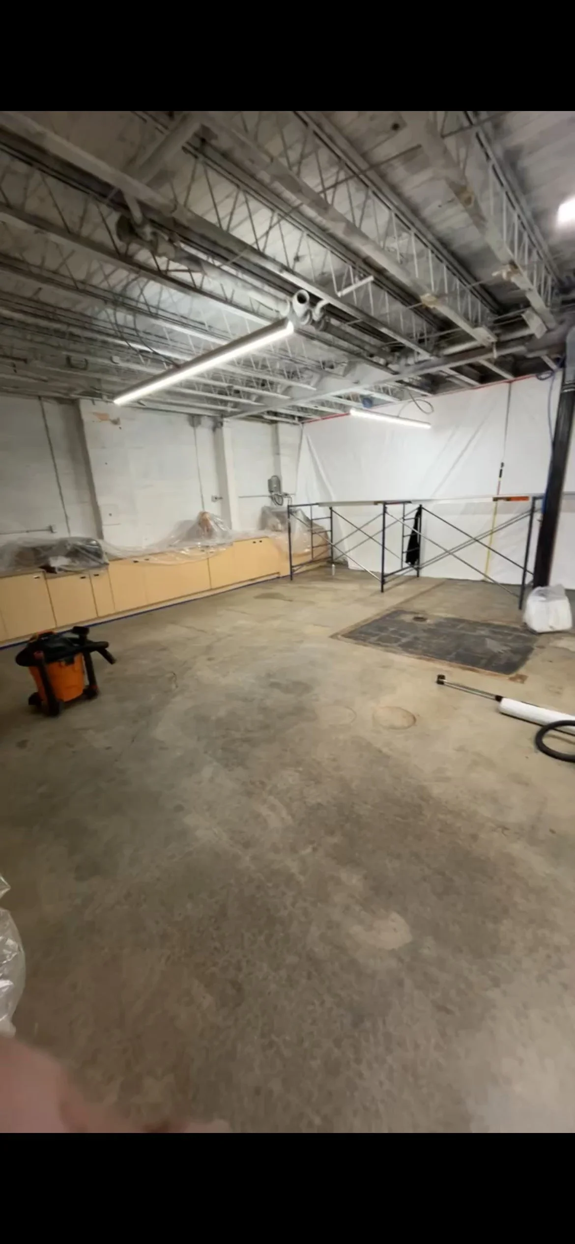 An unfinished interior space with a concrete floor, construction tools, scaffolding, and exposed ceiling pipes.