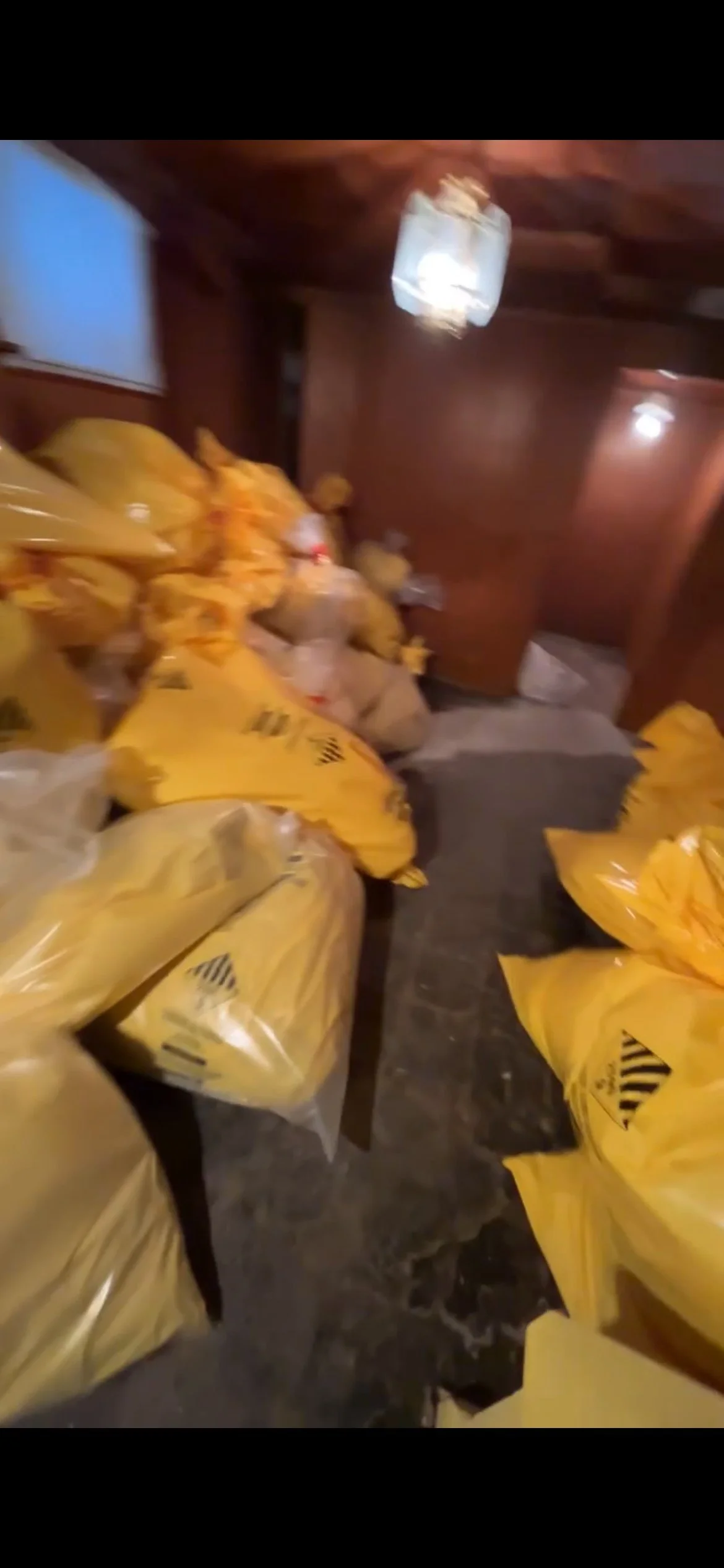 Room filled with numerous yellow plastic bags containing unknown items, on dark wooden tables and a gray carpeted floor, with a wooden wall and a ceiling light fixture.