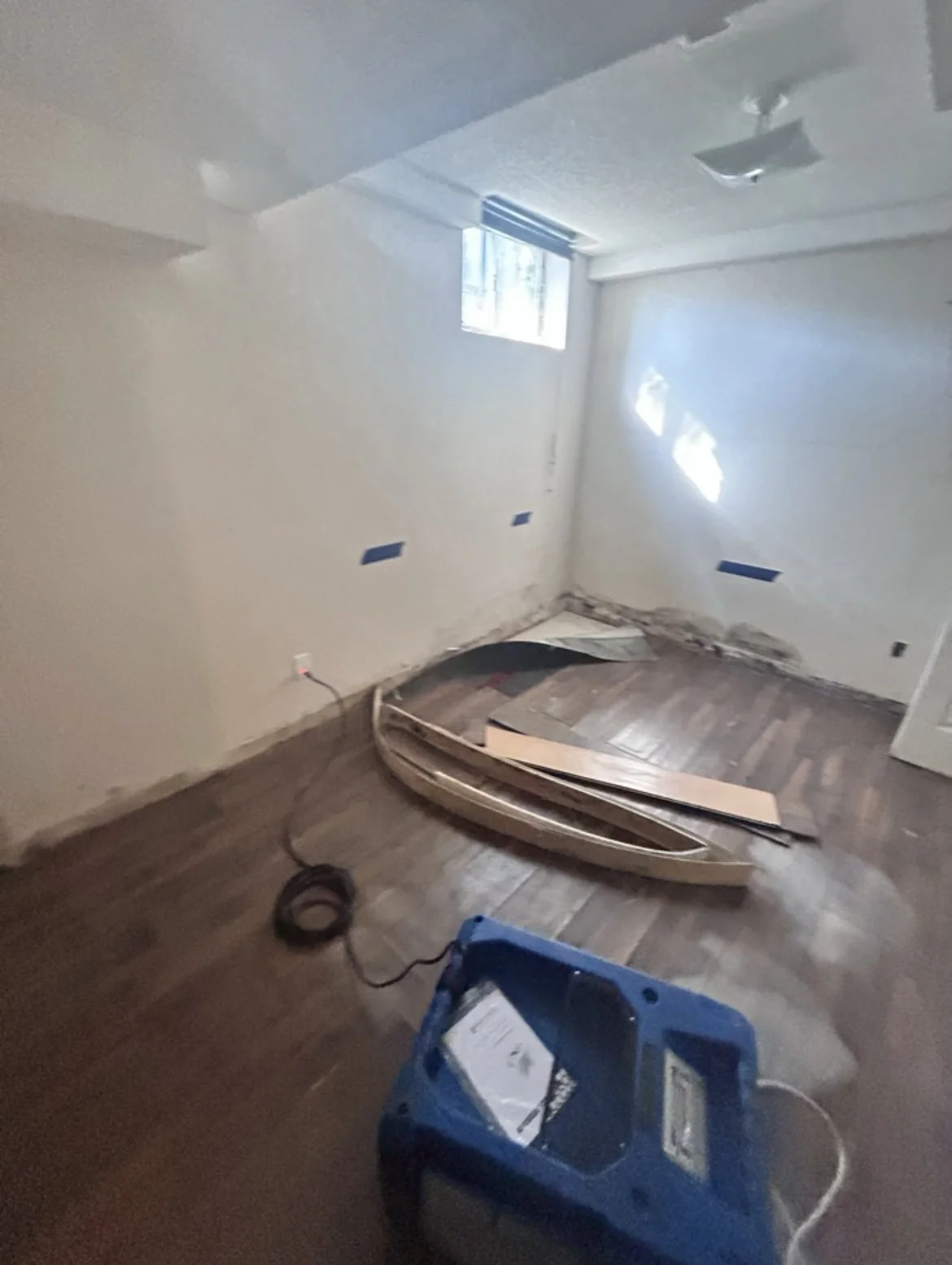 Room under renovation with a small boat, construction materials, and tools on the wooden floor, a window with blinds, and walls with electrical outlets and missing baseboards.