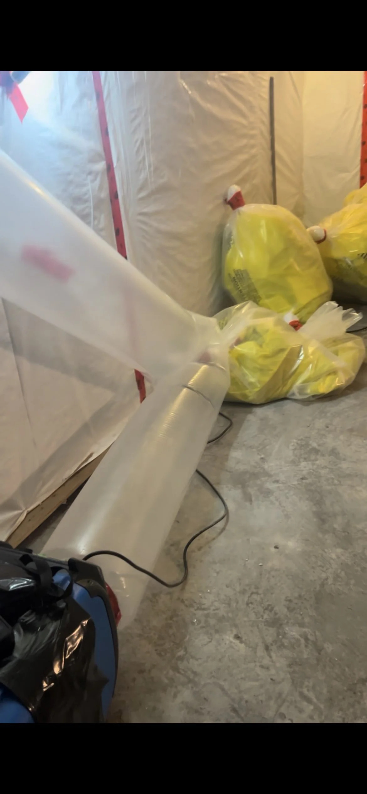 A space with plastic sheeting on the walls, yellow garbage bags filled with unknown items, and a heater on the floor connected with a power cord.