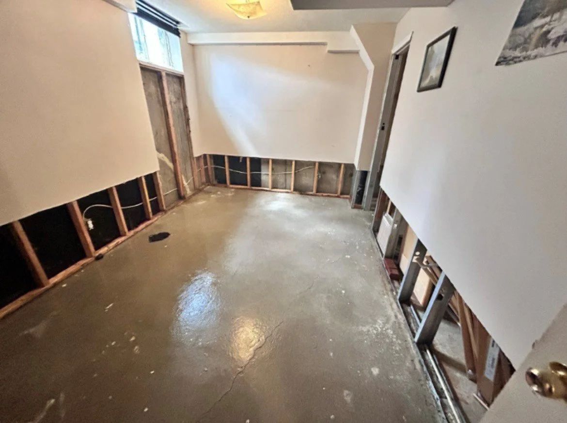 A room under renovation with exposed wall framing, a cement floor, partially finished walls, and a window on the left side.
