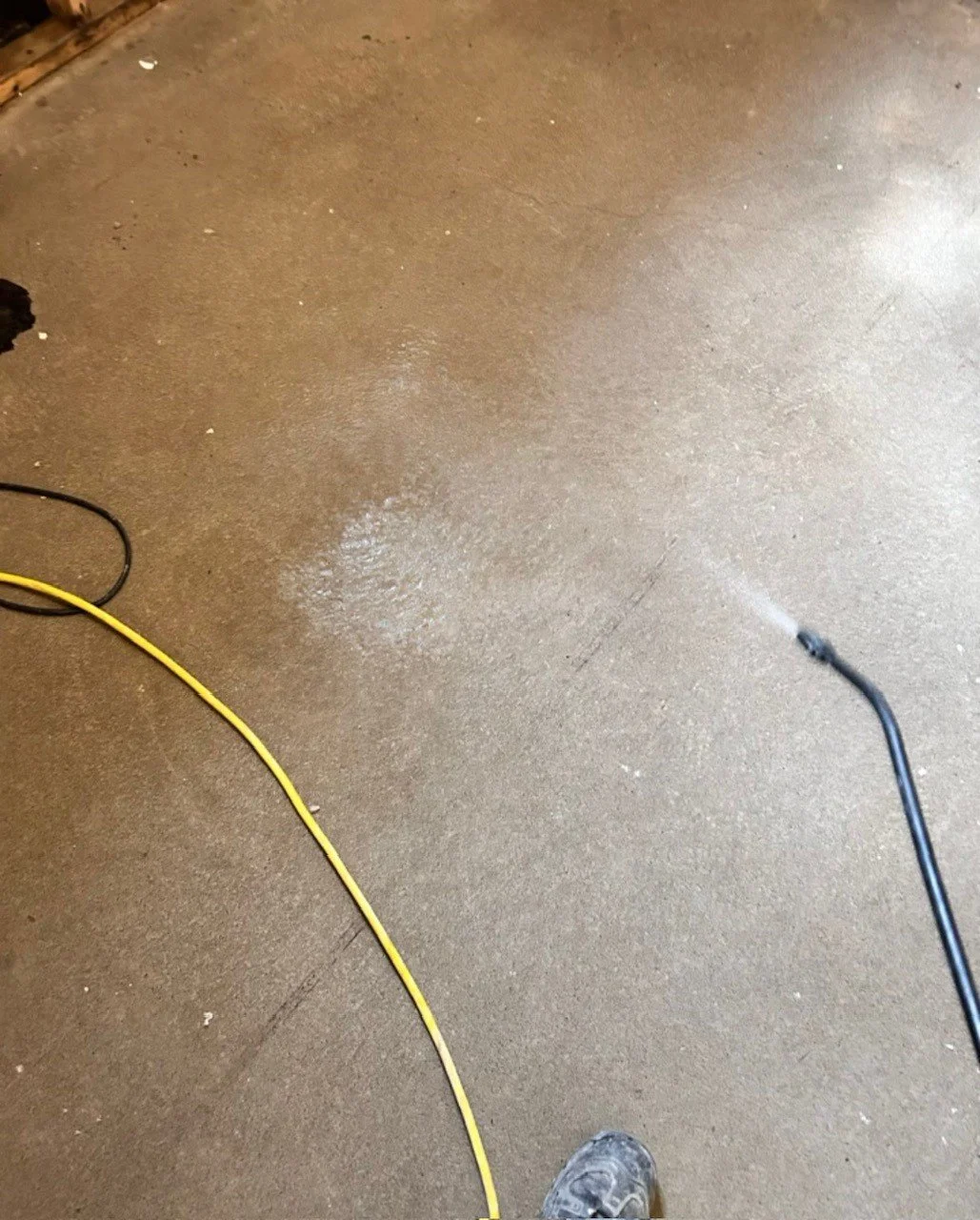 Person cleaning or spraying the floor with a pressure washer or spray wand, with yellow and black cords on a concrete floor.