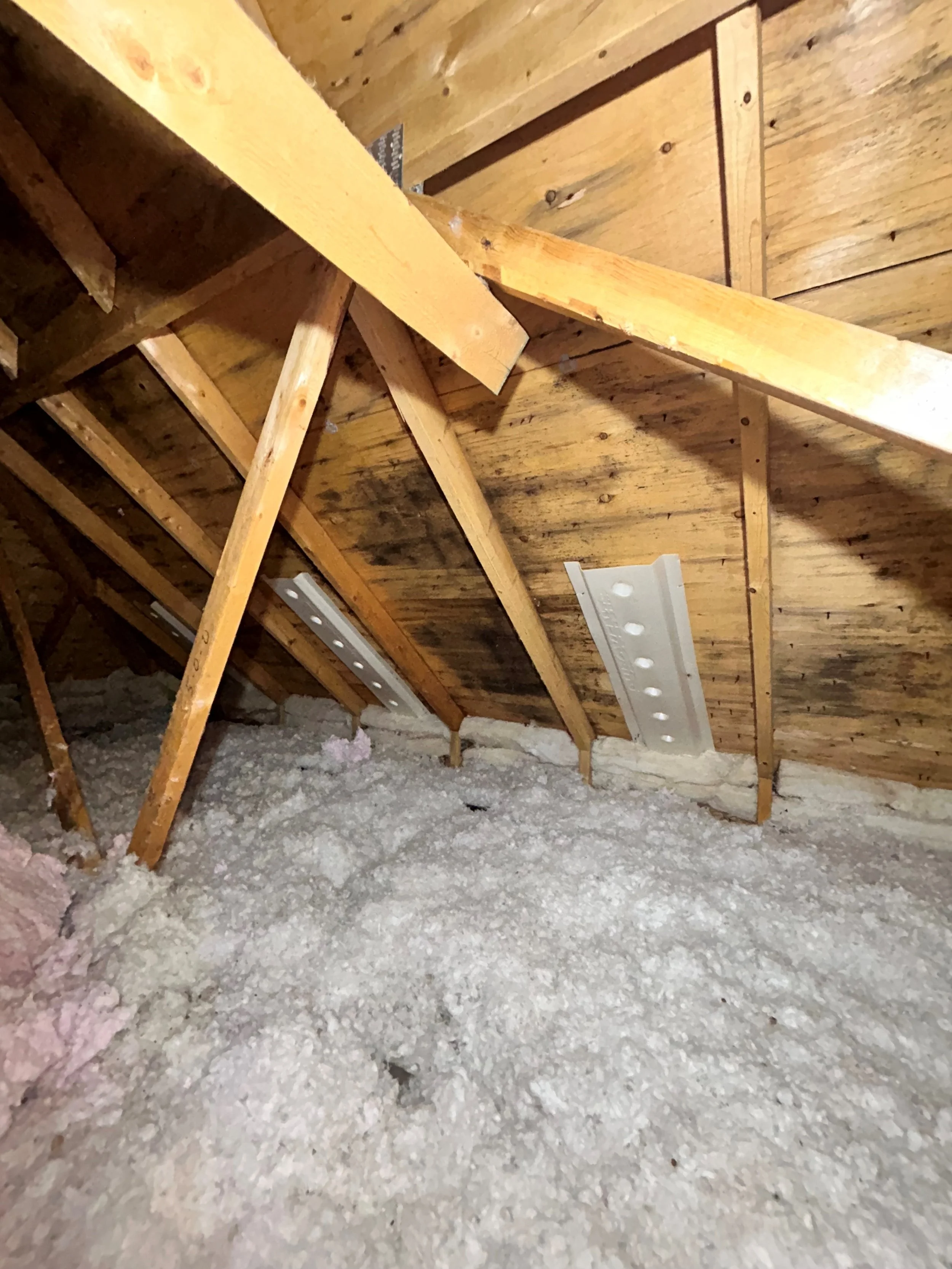 Attic with exposed wooden beams, spray foam insulation on the floor, and metal support brackets attached to the rafters.