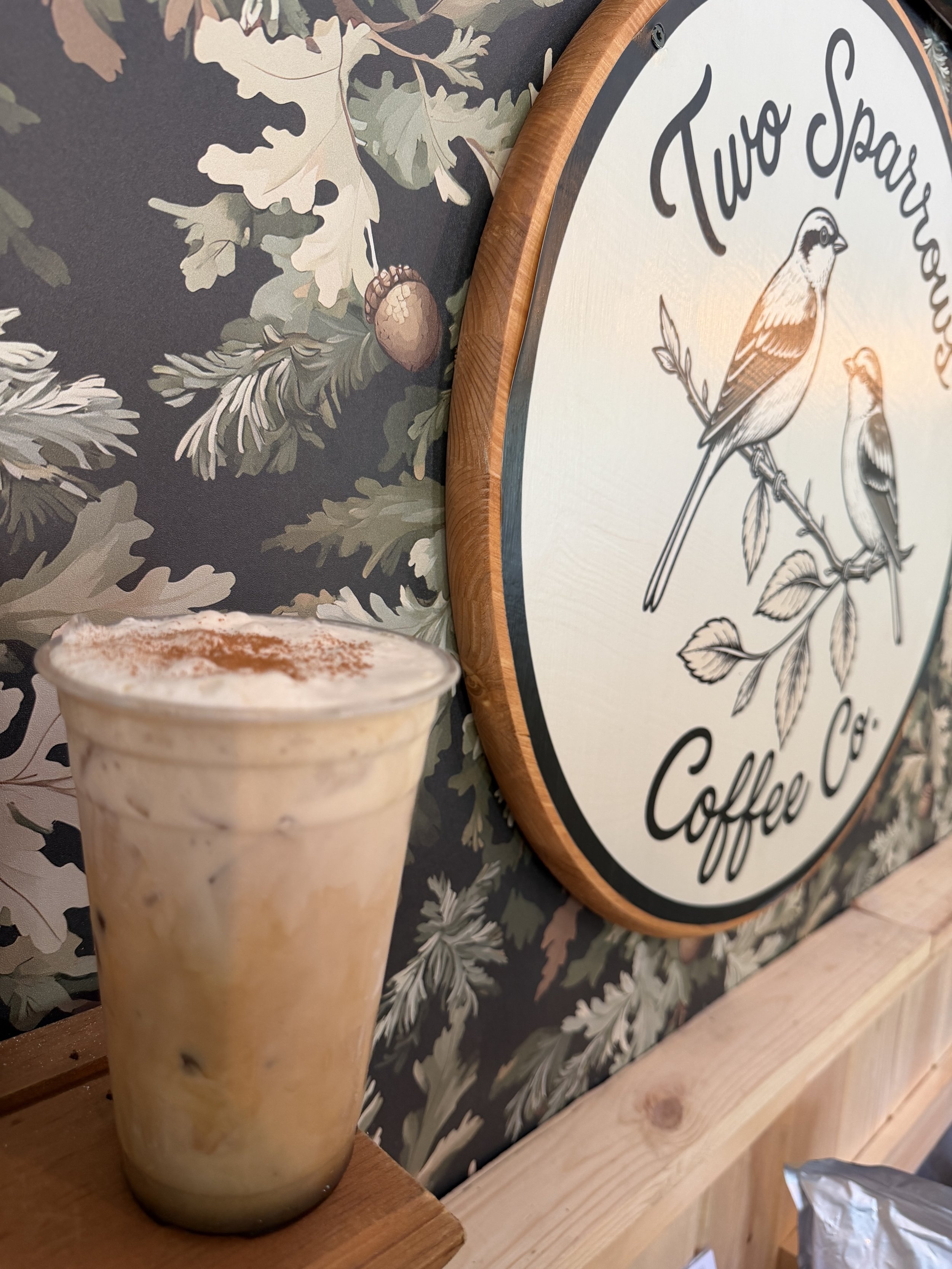 A creamy iced coffee sits on a ledge in front of a wall with dark wallpaper with acorns. There is a wood plaque with the Two Sparrows name and ink drawn sparrows.