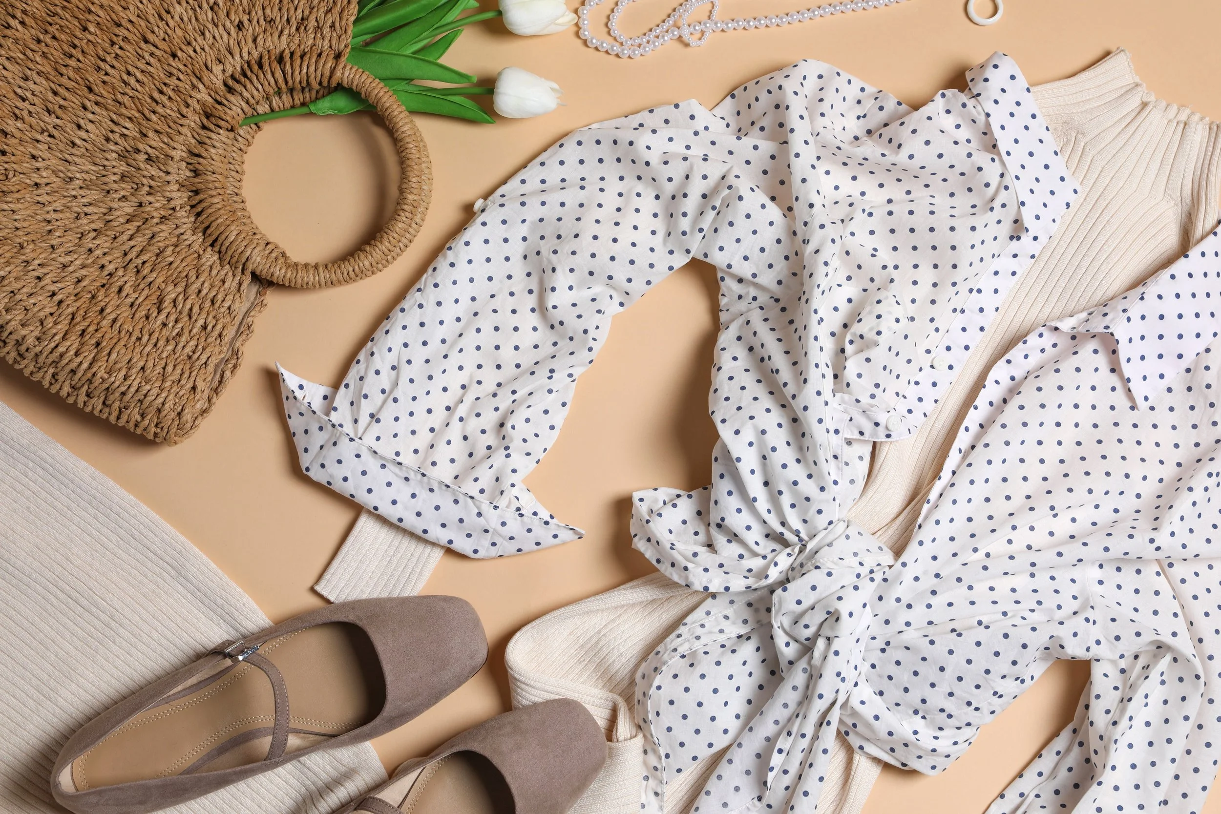 A flat lay of women's fashion and accessories, including a beige ribbed turtleneck sweater, a polka dot shirt with a tie-front detail, beige ankle boots, a woven straw handbag with circular handles, a pearl necklace, and white tulips with green leaves.