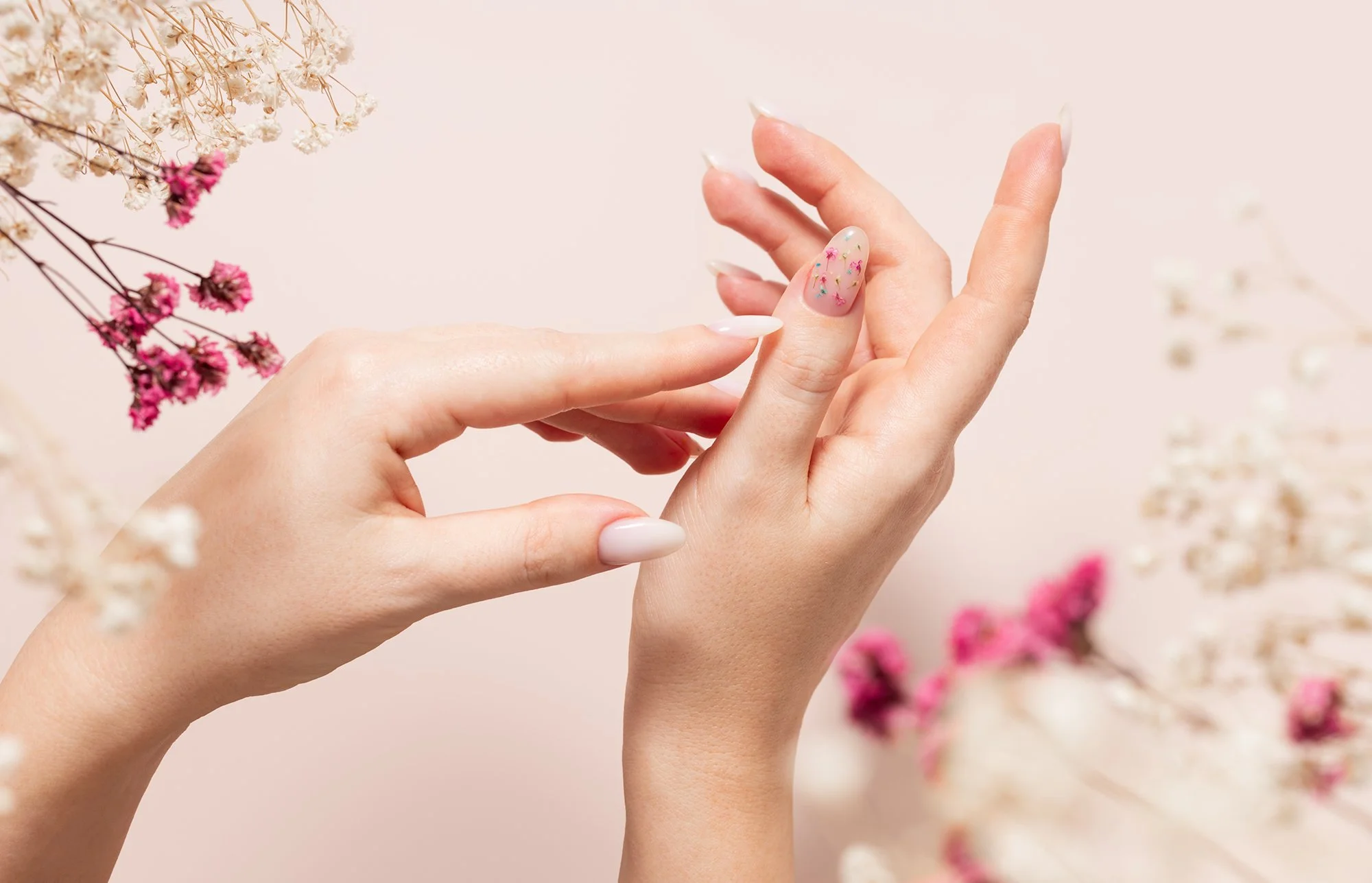 Close-up of hands with manicured nails, one hand gently touching the other, with delicate pink and white flowers in the background.