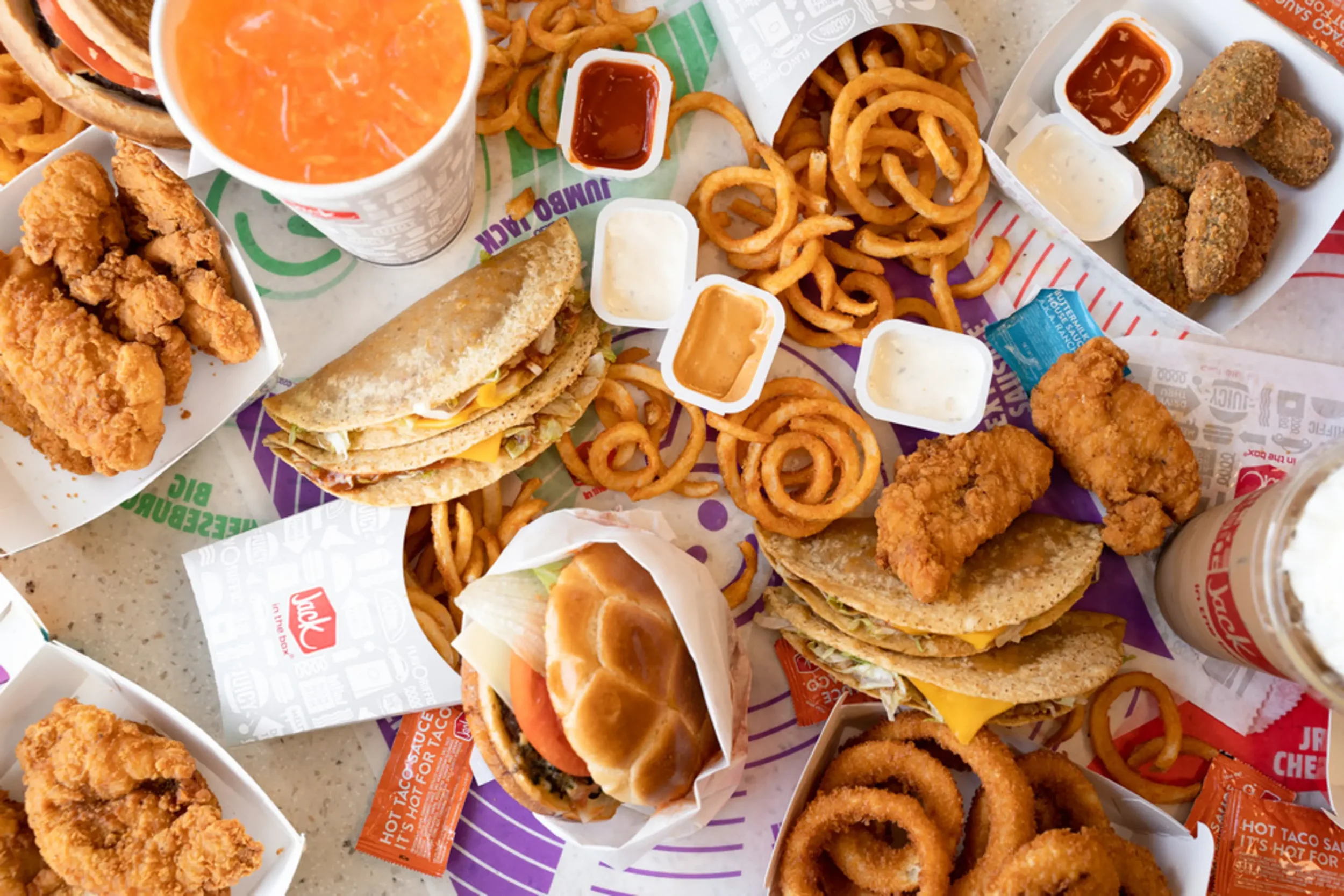 A table filled with fast food including chicken tenders, fried onion rings, curly fries, two cheeseburgers, a sandwich, and drinks, with multiple dipping sauces.