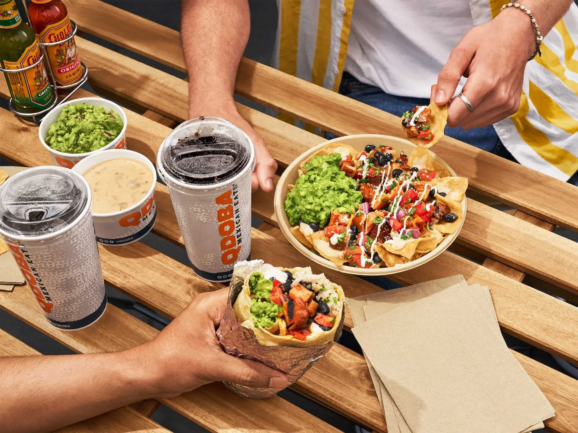 Two people sharing nachos with guacamole, salsa, and toppings, along with drinks, on a wooden picnic table.