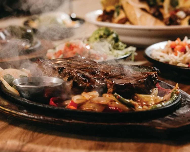 A sizzling plate with a cooked beef steak, sautéed vegetables, and a small container of sauce on a wooden table with sides of salad and chips in the background.