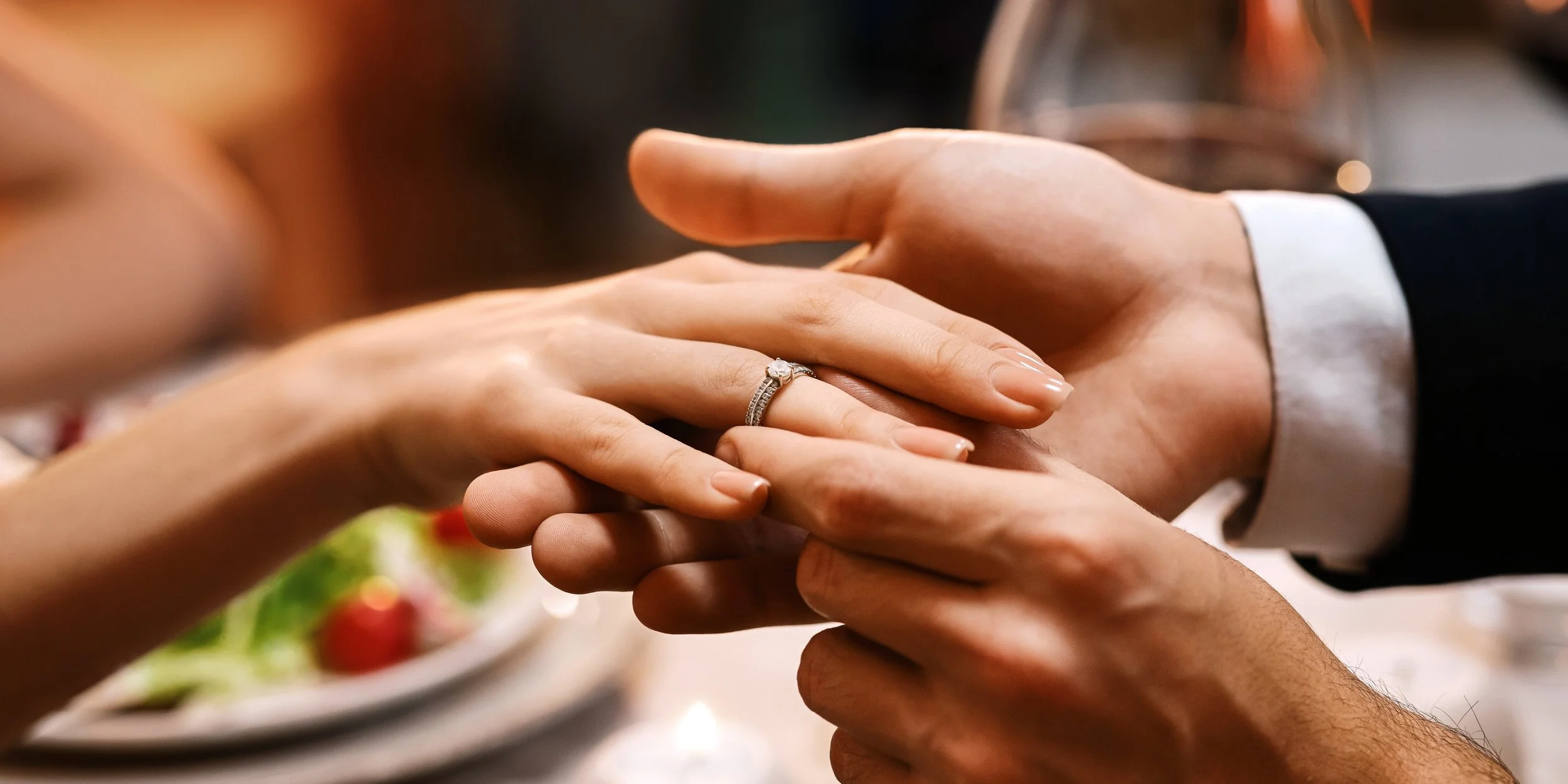 A close-up of a woman receiving a wedding or engagement ring from a man during a ceremony. The woman has a diamond ring on her finger, and the man's hand is gently holding hers.