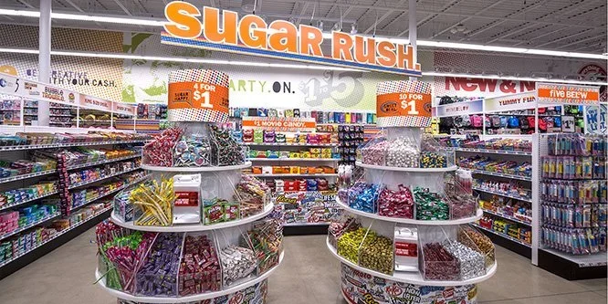 Candy aisle in a store with circular displays of assorted candies and a large orange and white Sugar Rush sign overhead.