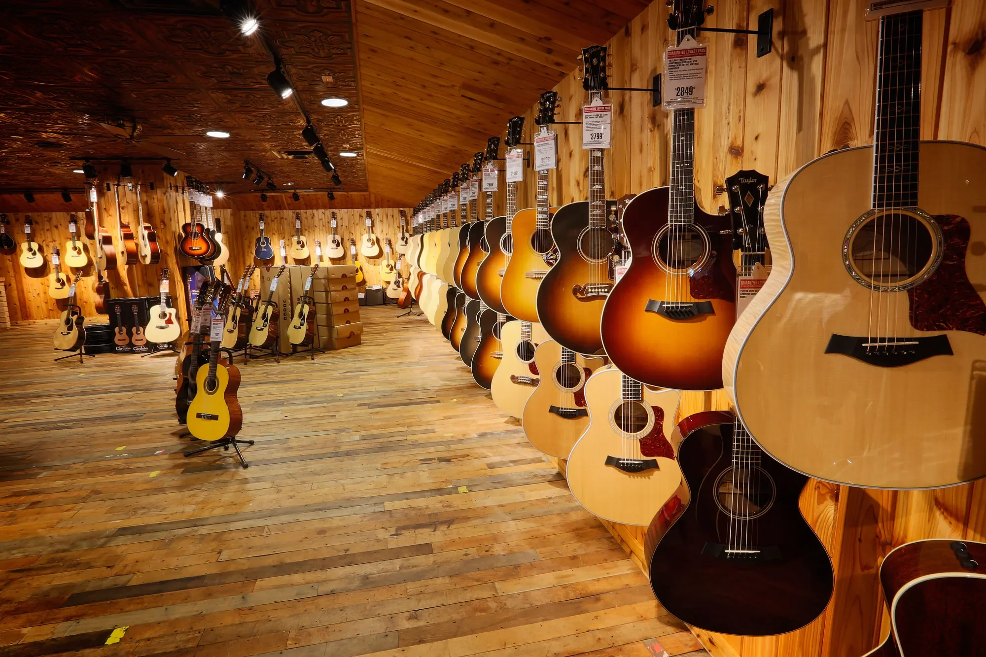 A guitar shop with wooden walls and floors, displaying a variety of acoustic guitars on the wall and on stands, with some guitars hanging from the ceiling.