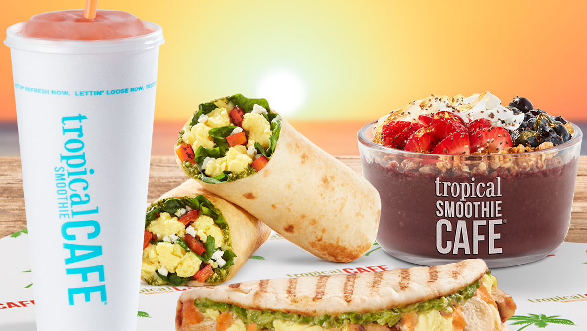 A meal from Tropical Smoothie Cafe including a smoothie, a wrap, a sandwich, and a smoothie bowl on a wooden table with a sunset background.