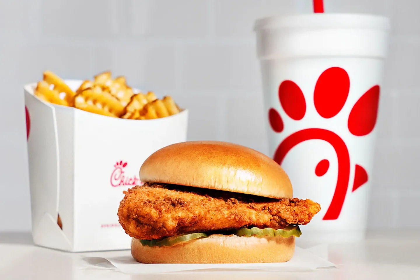 Chick-fil-A fried chicken sandwich with lettuce and pickles, a side of waffle fries in an open Chick-fil-A box, and a Chick-fil-A drink cup with a red logo.