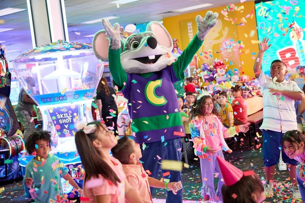 Children at a birthday party with a mascot dressed as a colorful mouse, confetti falling, and arcade games in the background.