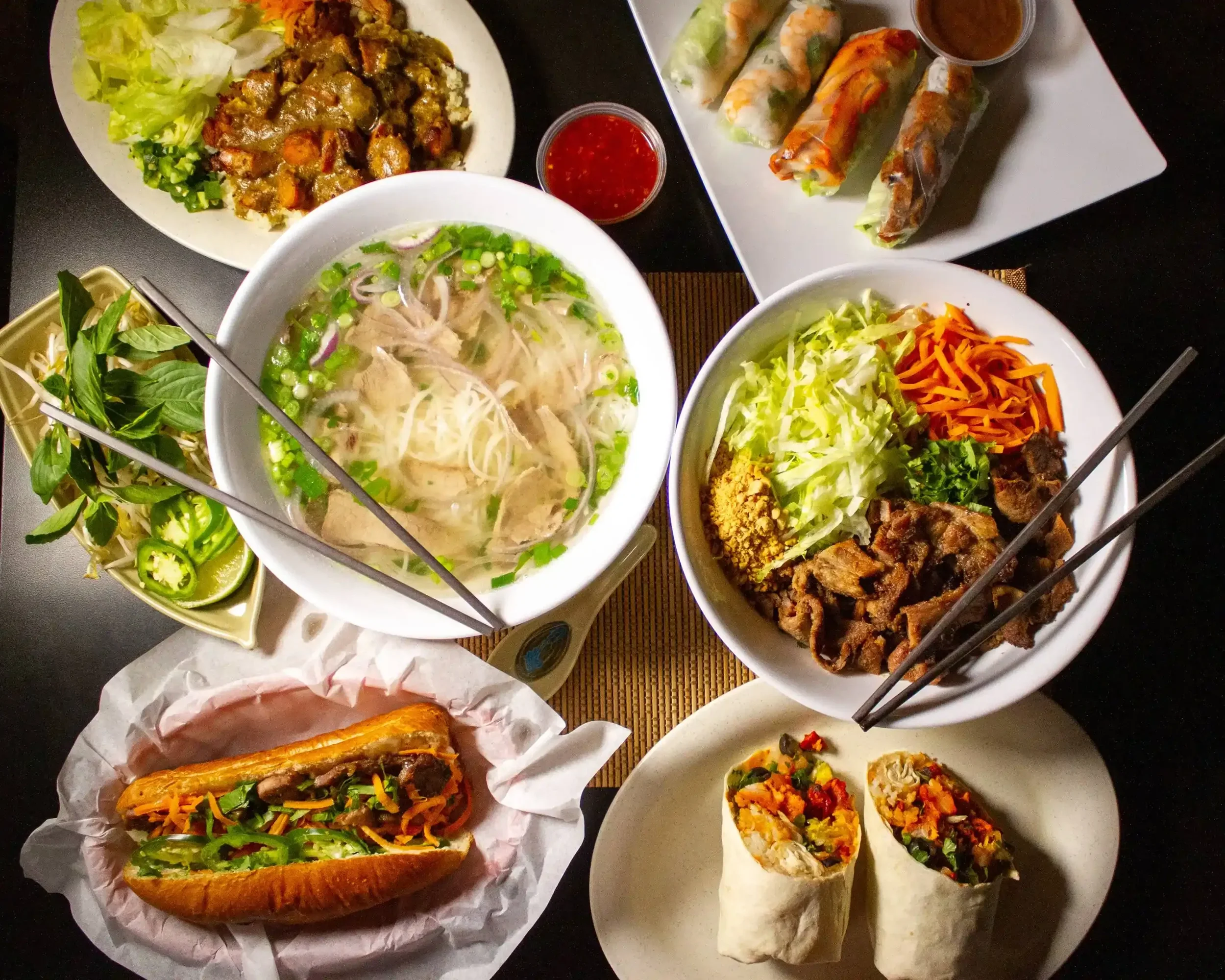 A variety of Vietnamese dishes on a table, including a bowl of Pho with shredded chicken and green onions, spring rolls with dipping sauce, a bowl of grilled meat with vegetables and cover, a banh mi sandwich with vegetables and meat, and two vegetable spring rolls.
