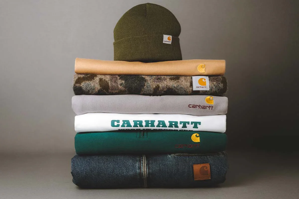 Stacked clothing including a green beanie, beige fabric, camouflage shirt, gray sweatshirt, white Carhartt T-shirt, green Carhartt hoodie, and dark gray Carhartt jacket on a flat surface.