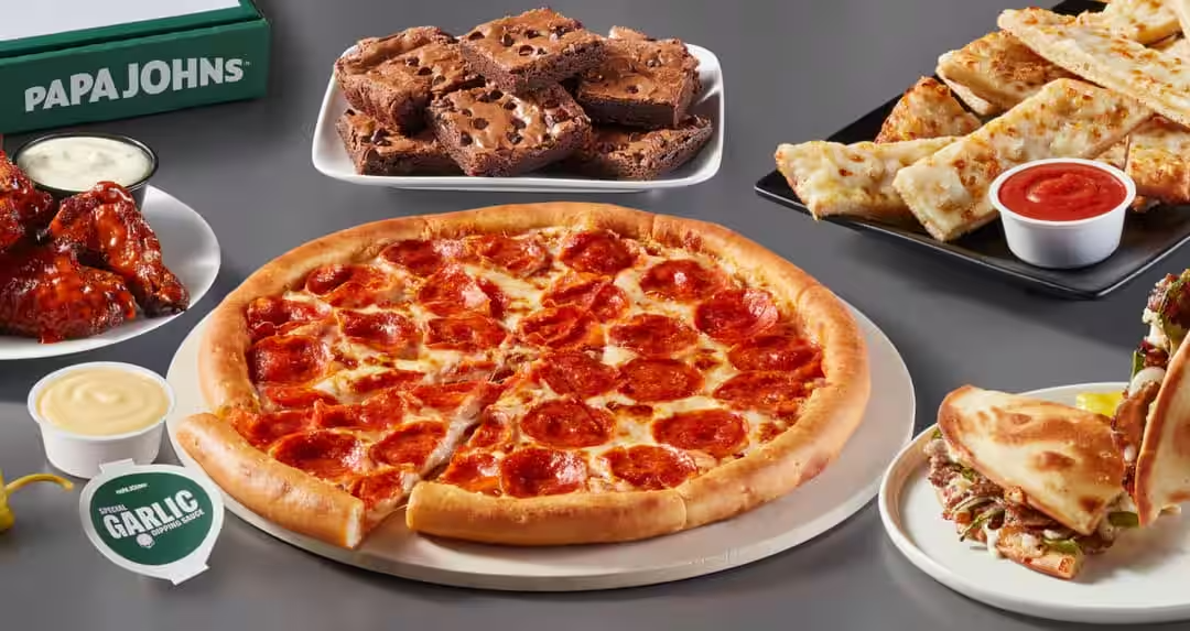 A pepperoni pizza with crust, garlic sauce, breadsticks with marinara, chicken wings with ranch, and a sandwich with sliced meat and vegetables on plates.