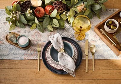 A table setting with a black plate, a cloth napkin, a fork, knife, and spoon, and a centerpiece of greenery and flowers. There is a glass of white wine and small bowls of olives and salt on the table.
