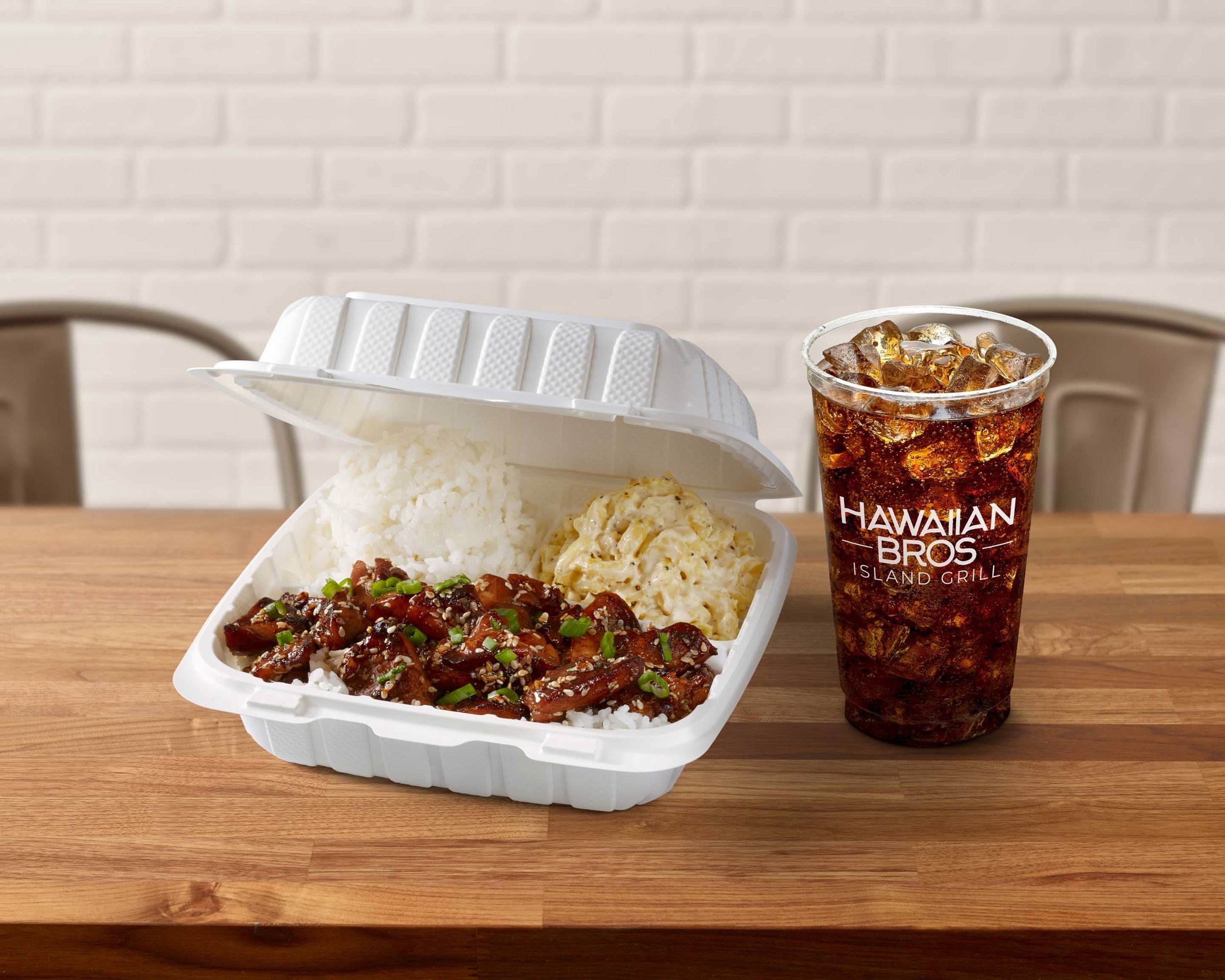 Hawaiian bros takeout meal with rice, macaroni salad, and teriyaki chicken, alongside a soda in a Hawaiian Bros Island Grill cup on a wooden table.