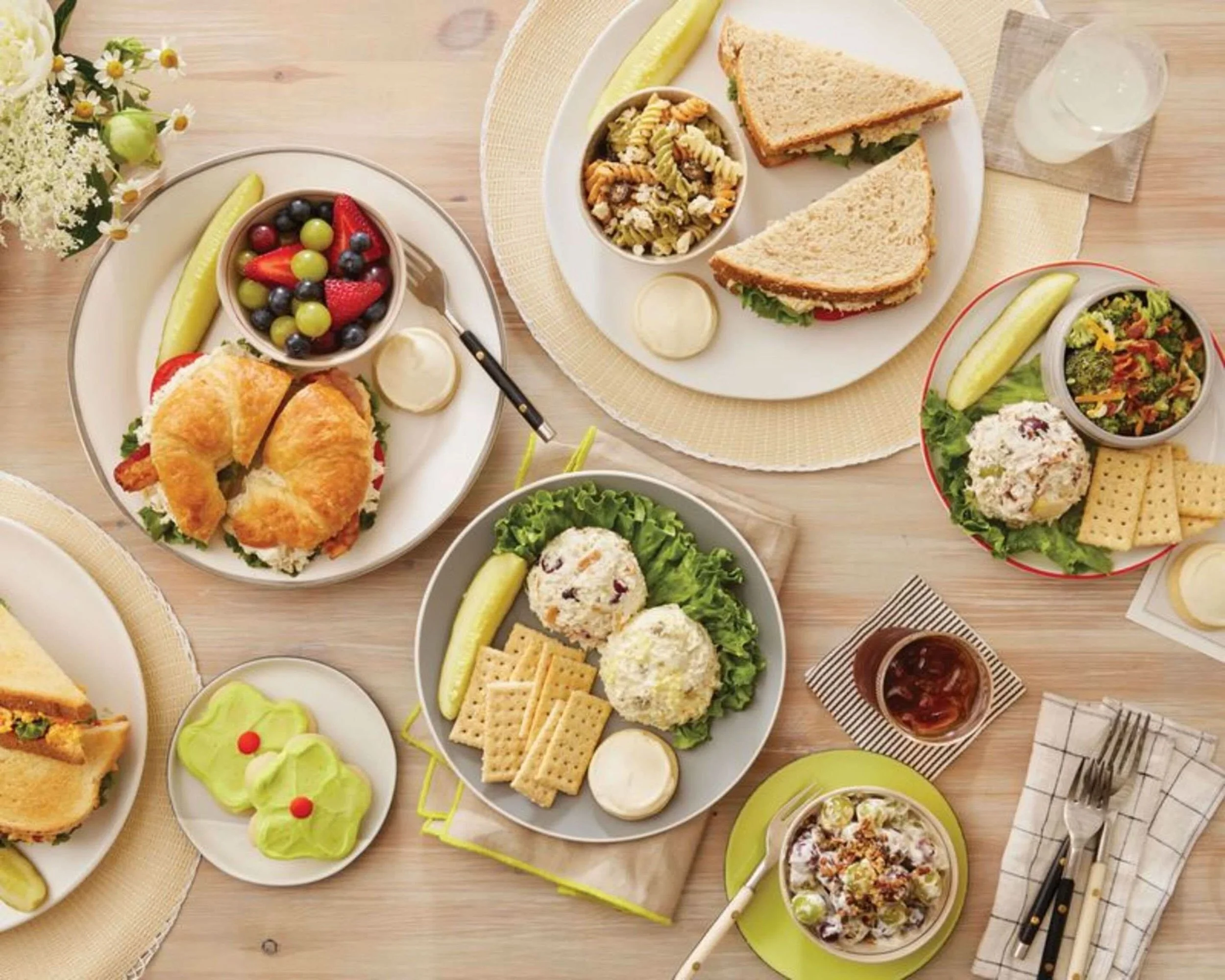 A variety of dishes on a wooden table, including sandwiches, salads, fruit, cookies, and beverages, arranged on different plates and bowls.