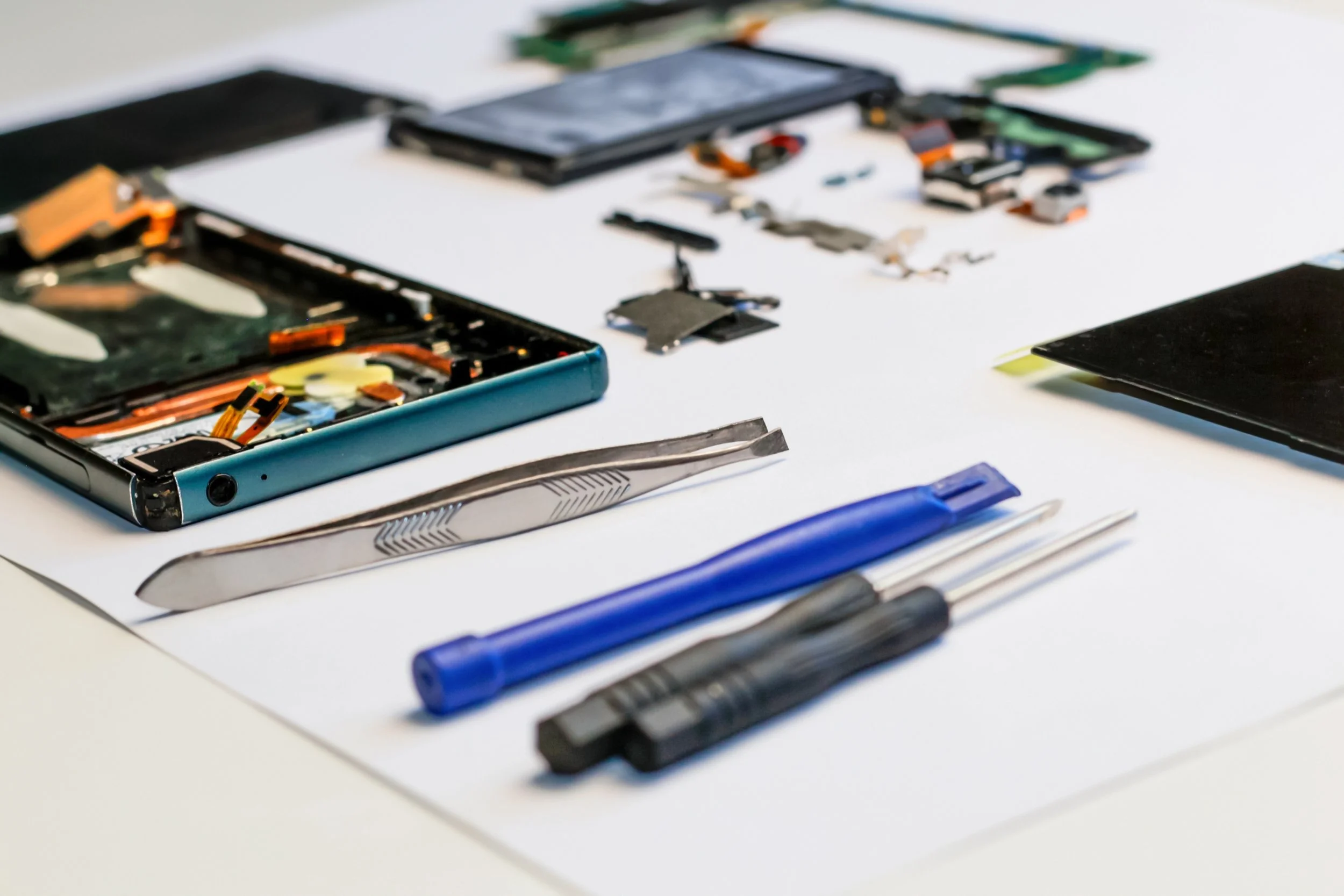 Disassembled smartphone with various tools including plastic opening tools, tweezers, screwdrivers, and small components laid out on a white surface.