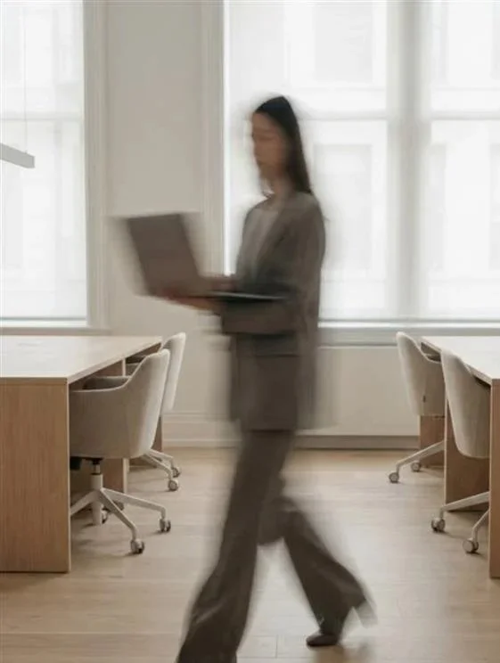 A blurred woman walks in an office space with white walls, wooden desks, and beige chairs, holding a laptop.