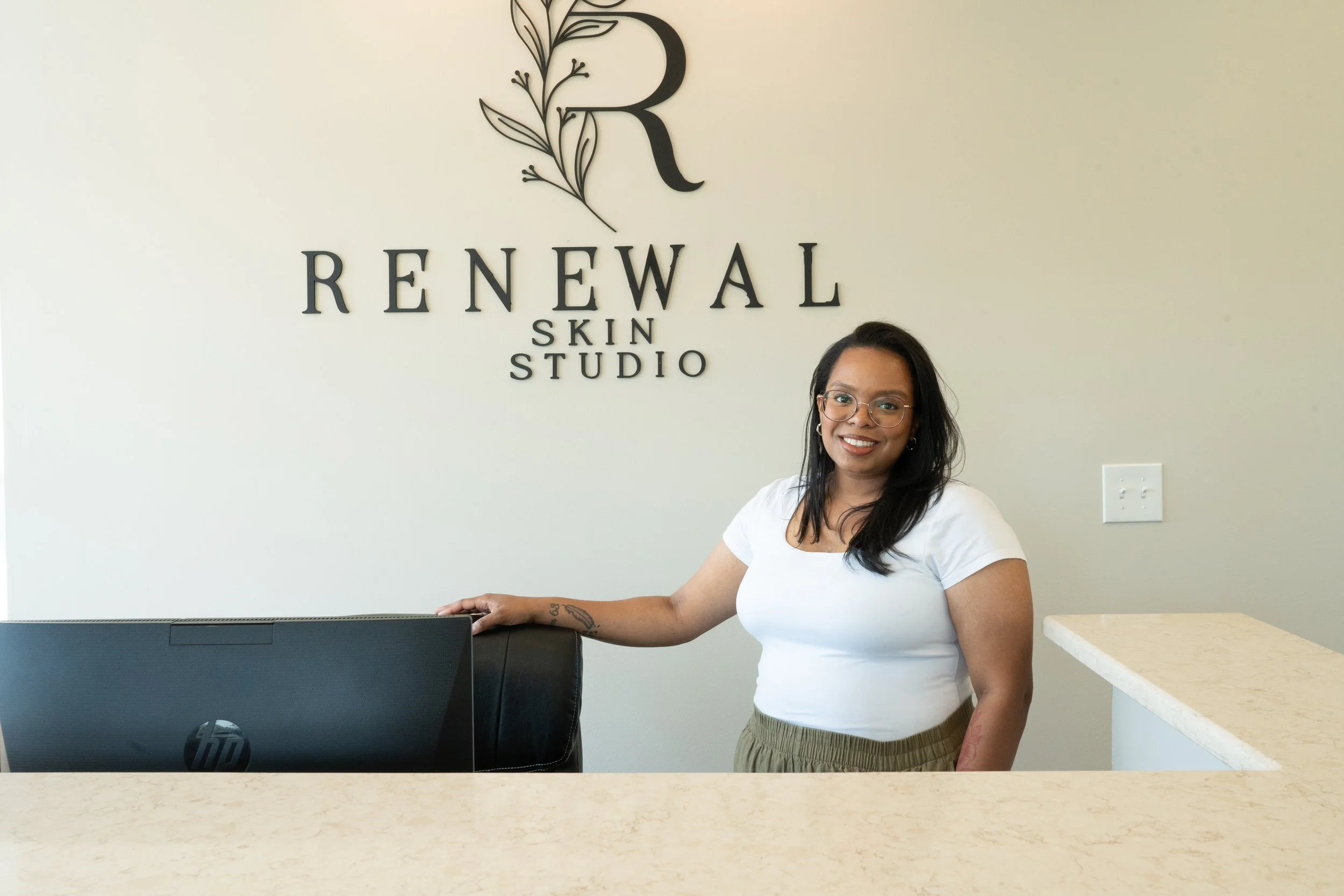 A woman with glasses and a white shirt standing behind a reception desk in a skin studio called Renewal Skin Studio, smiling at the camera.