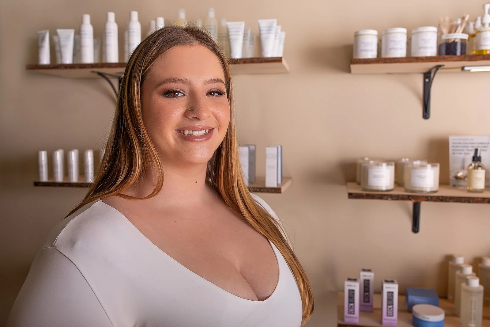 A smiling woman with long red hair wearing a white top standing in front of shelves filled with skincare or cosmetic products in a store or spa.