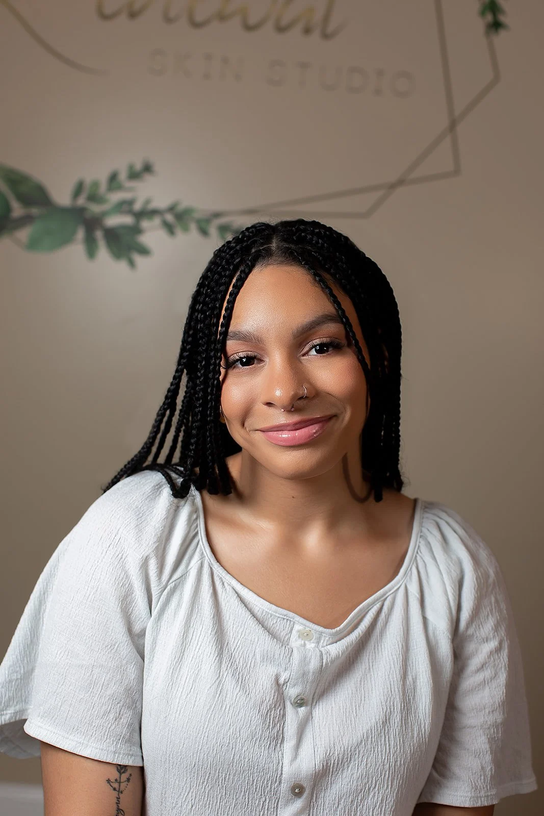 A young woman with medium dark skin, long braided hair, and a small nose ring, smiling at the camera. She is wearing a light-colored button-up shirt. The background features a wall with the words 'Creative Skin Studio' and some green leaf graphics.