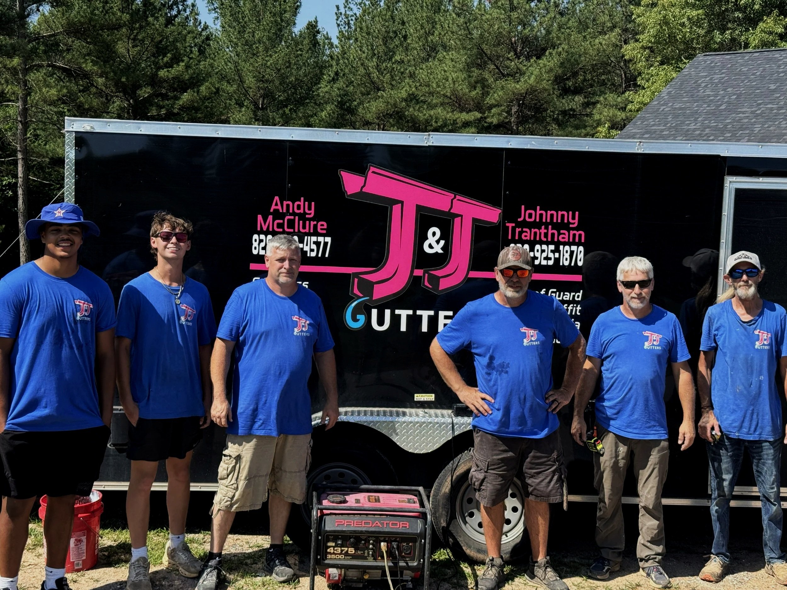 Group of people standing outdoors in front of a black trailer. They are wearing blue T-shirts with a logo. Some are smiling and wearing sunglasses or hats. There is a portable generator on the ground in front of them.