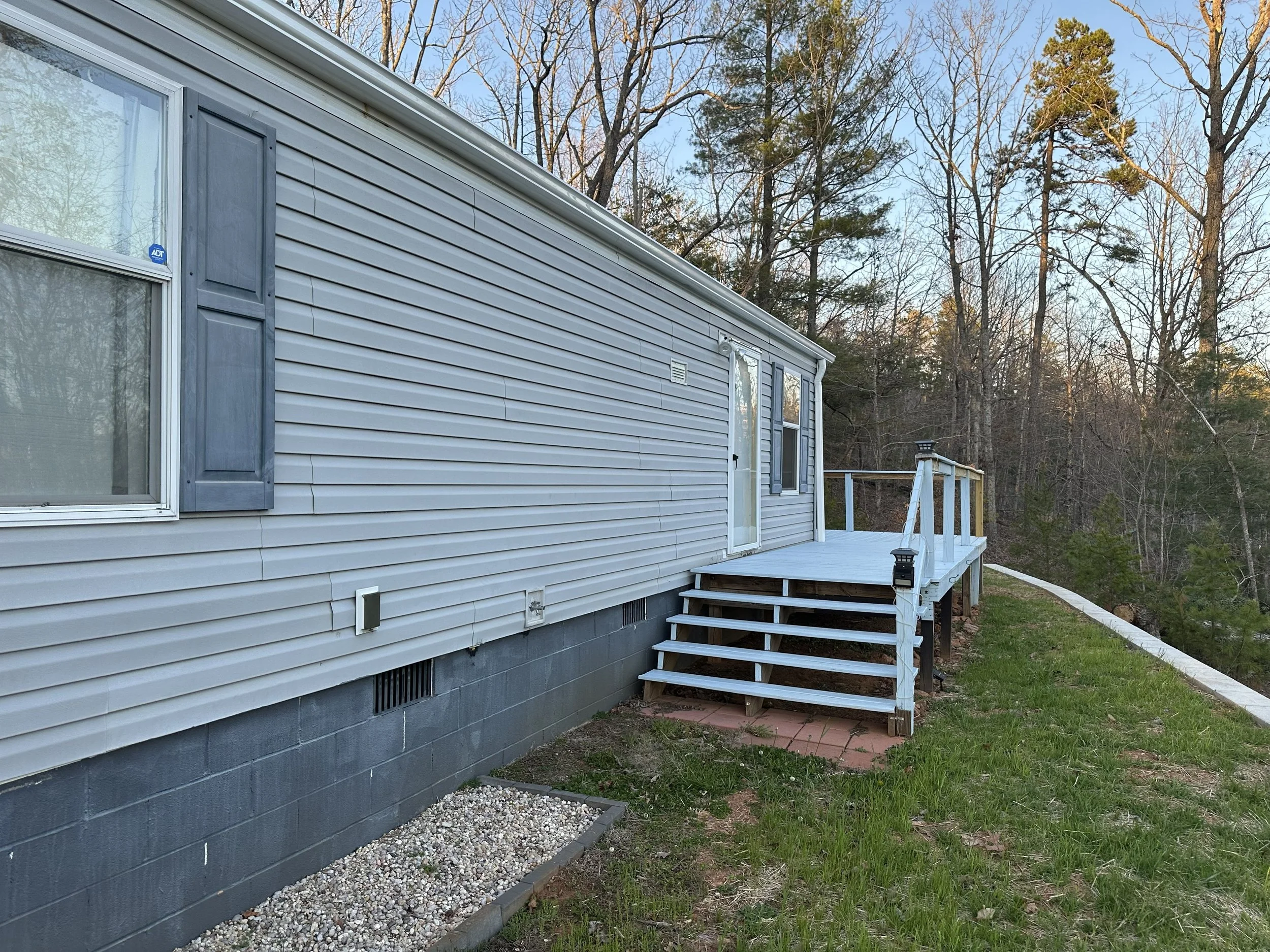 Side view of a house with gray siding on a hillside, with a small deck, steps, and a yard with grass and trees in the background.