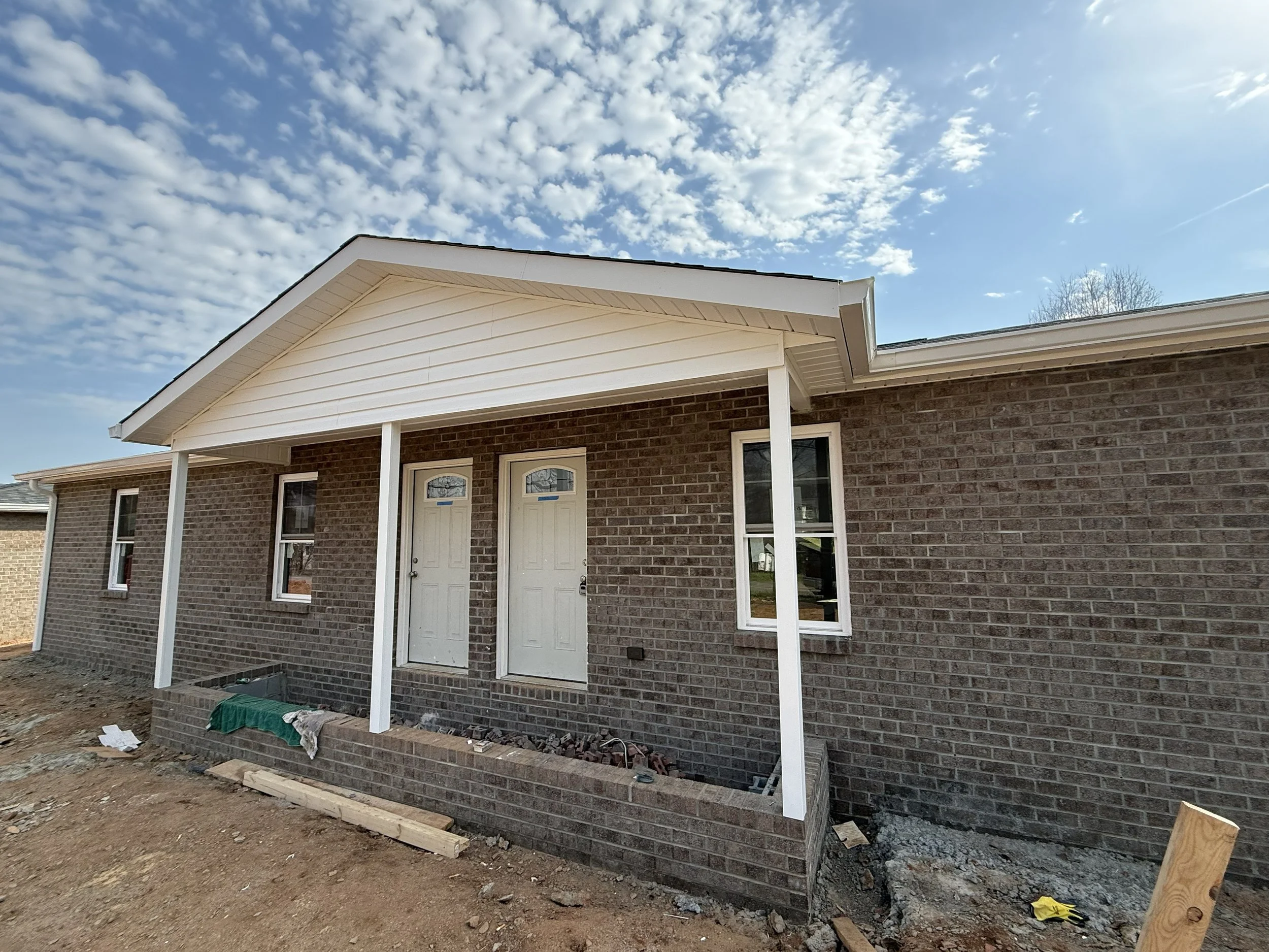 Front of a brick house under construction with a porch, white trim, and a partly finished yard.