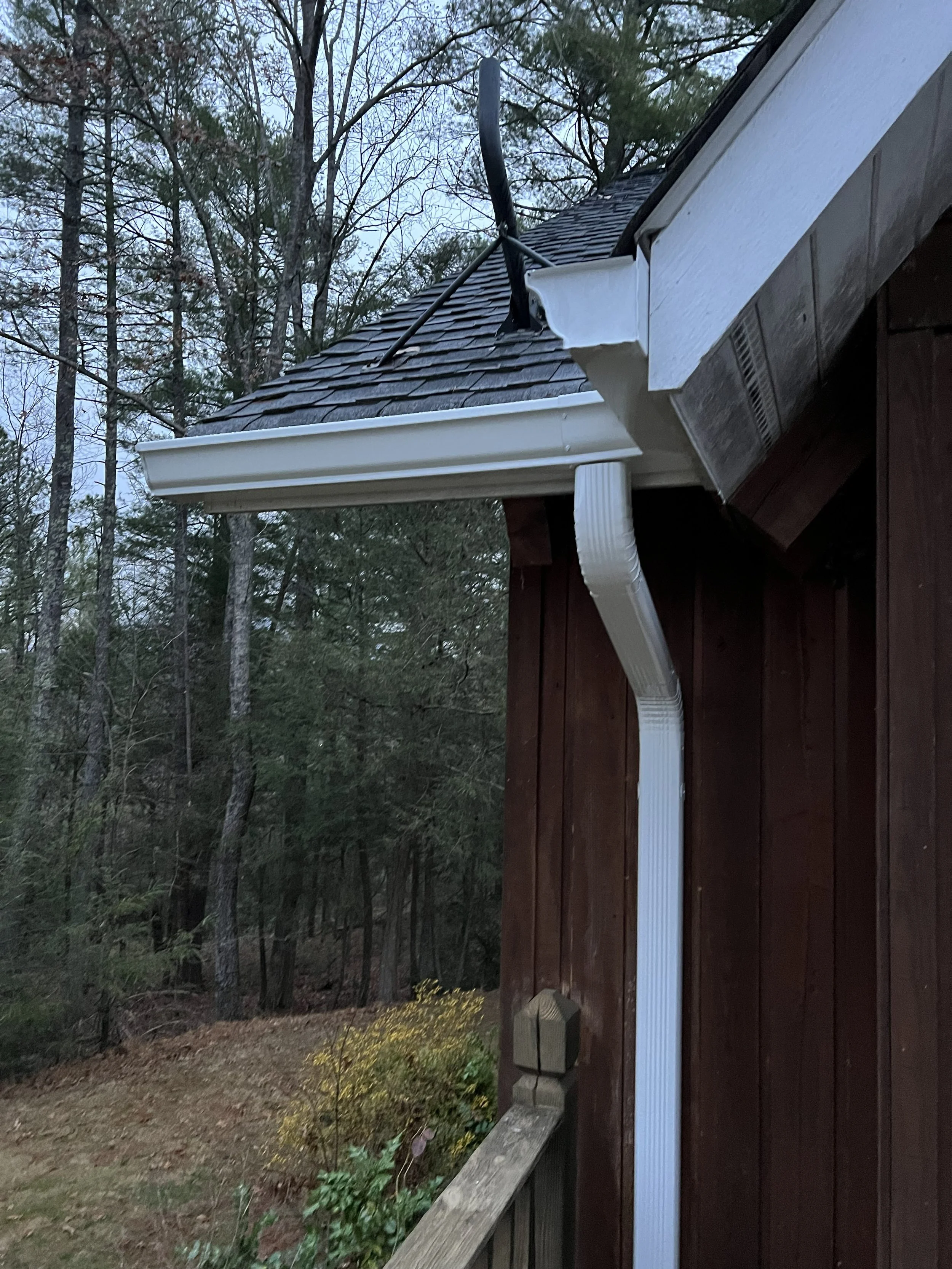 Side of a house with a sagging black antenna and a white rain gutter, set against a background of trees with sparse leaves, indicating fall or early winter.