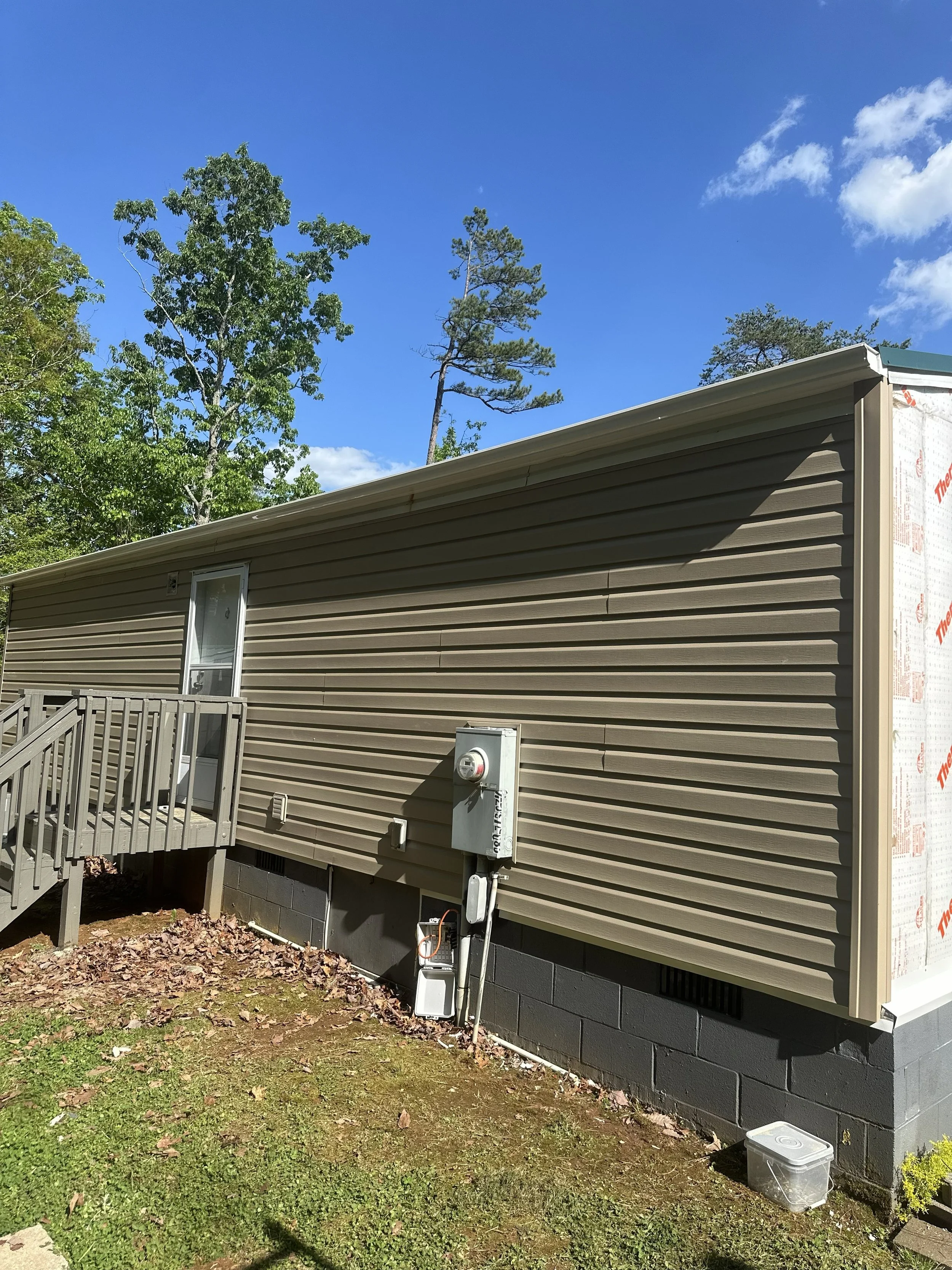 Side view of a beige vinyl-sided house with a wooden deck and stairs, electrical meters, and a plastic storage container outside, surrounded by grass and trees under a blue sky with a few clouds.