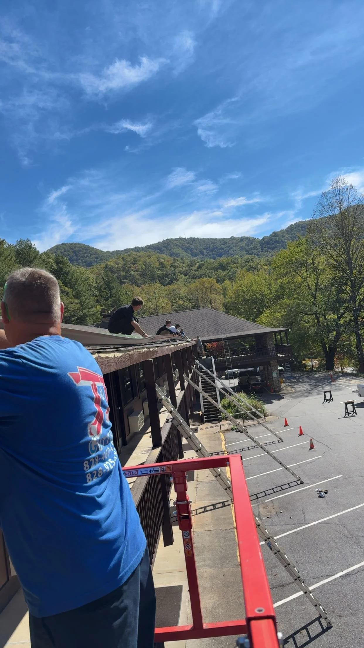 People working on the roof of a building with ladders and scaffolding, with a backdrop of green forested mountains and a partly cloudy sky.