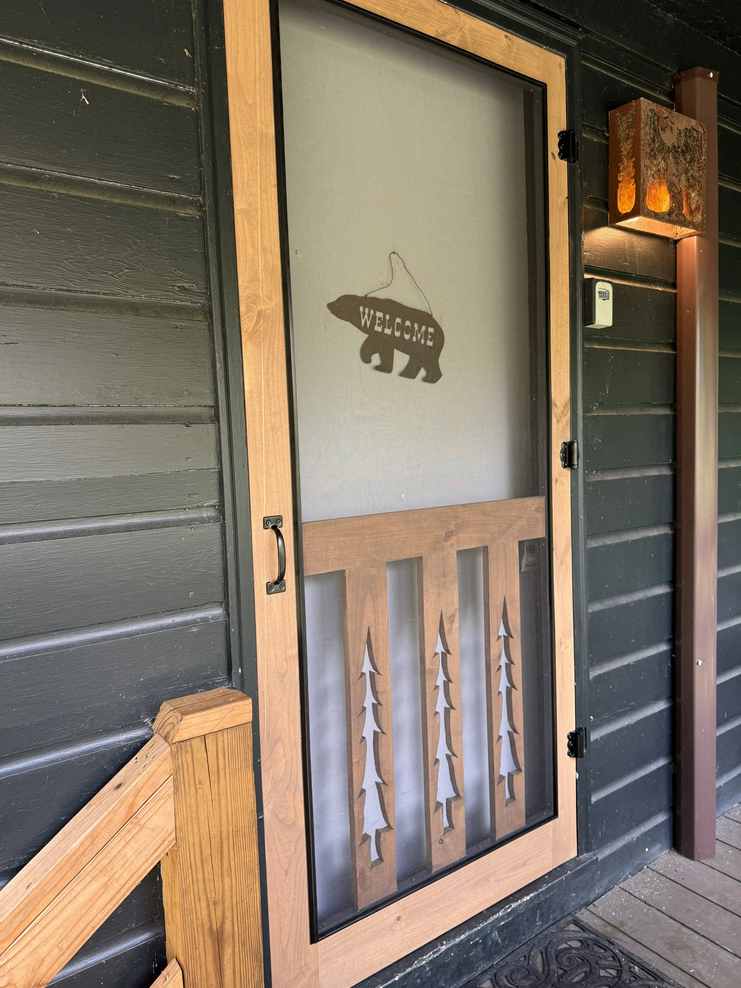 A screen door with a bear silhouette and the word 'WELCOME' on a piece of fabric, mounted on a wooden frame, outside a dark-painted building.