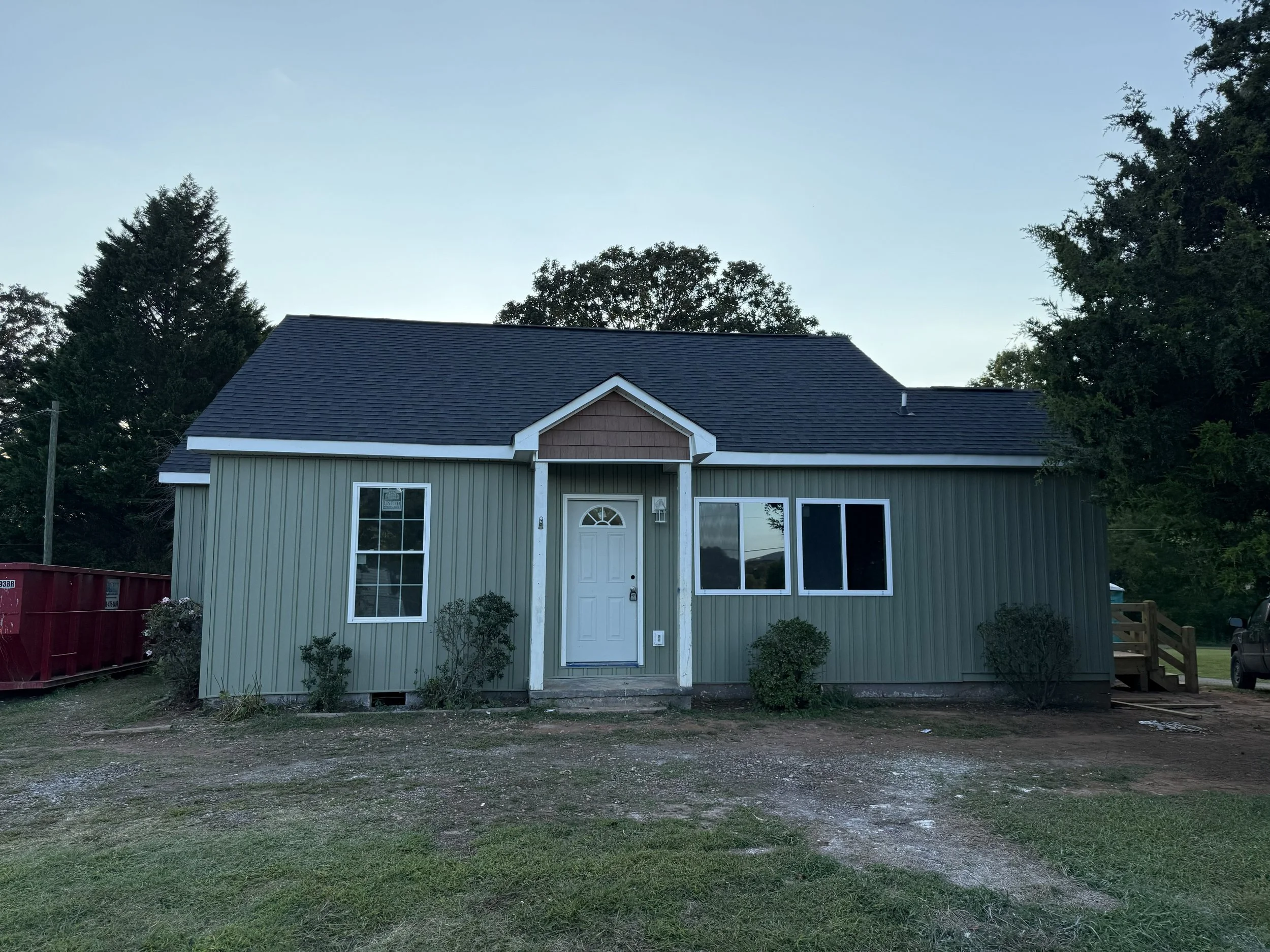 A house with a green exterior, a gabled roof, and a front porch, surrounded by trees and vegetation.