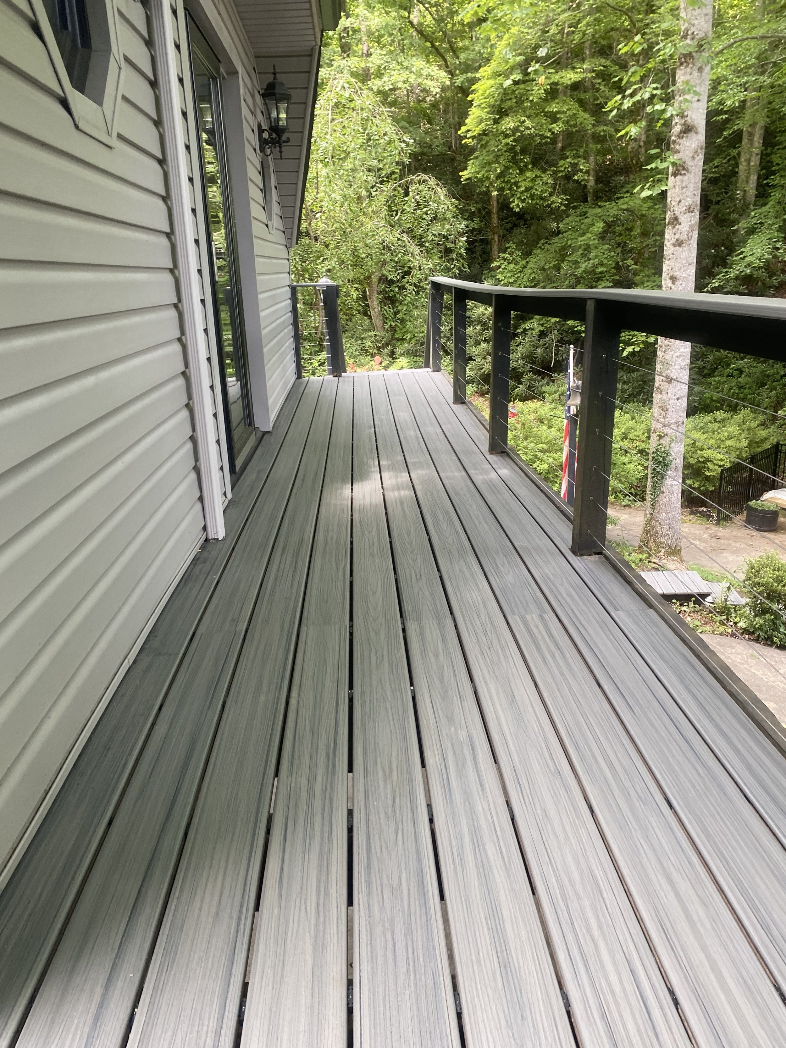 A wooden balcony with black railing and wiring, attached to a house with beige horizontal siding, overlooking a green forest with trees and sunlight filtering through.