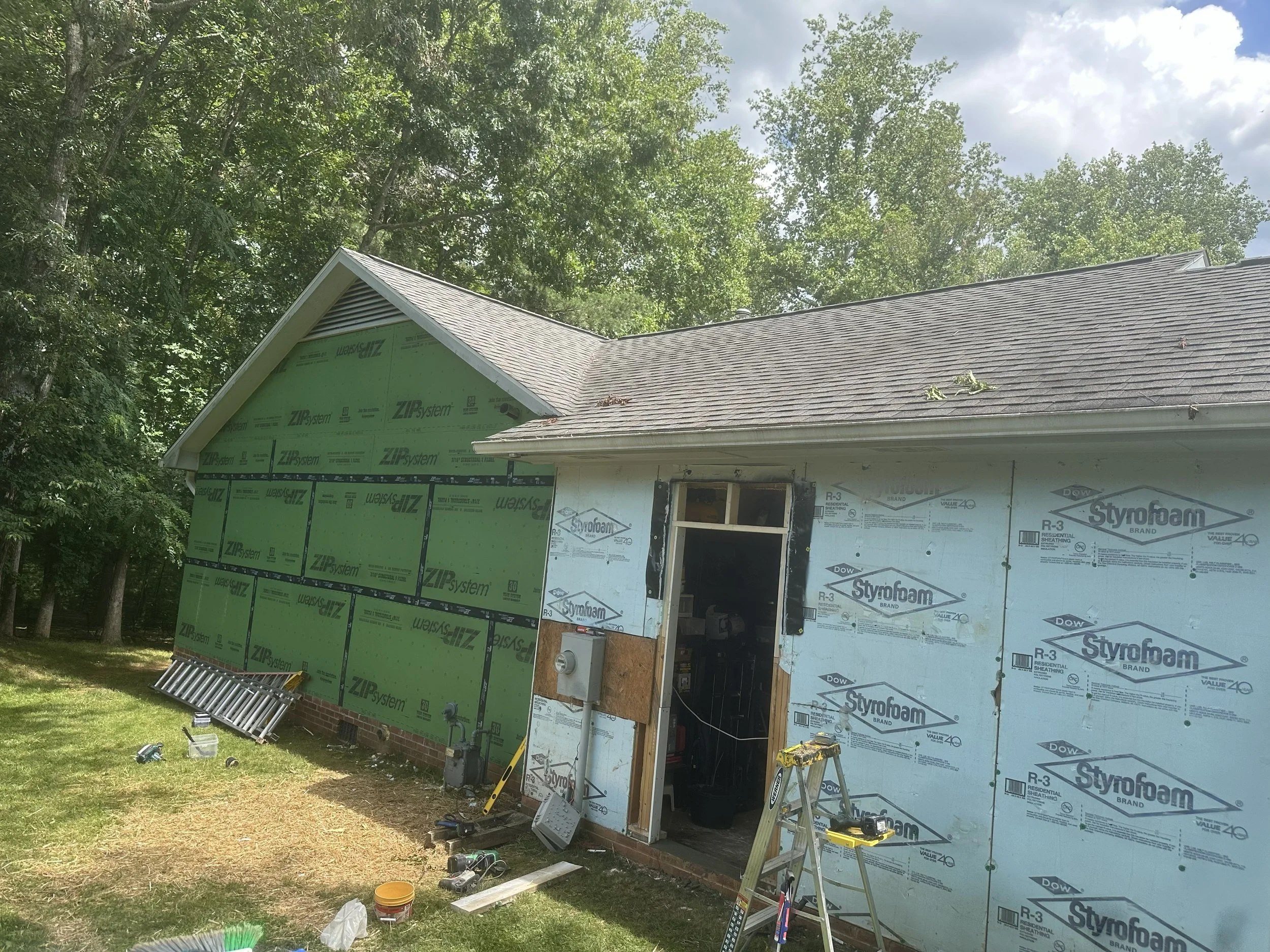A house under construction with partially finished walls covered in insulation and sheathing. Construction tools and materials are scattered in the yard, and the roof has roofing shingles installed. There are large trees in the background and a partl