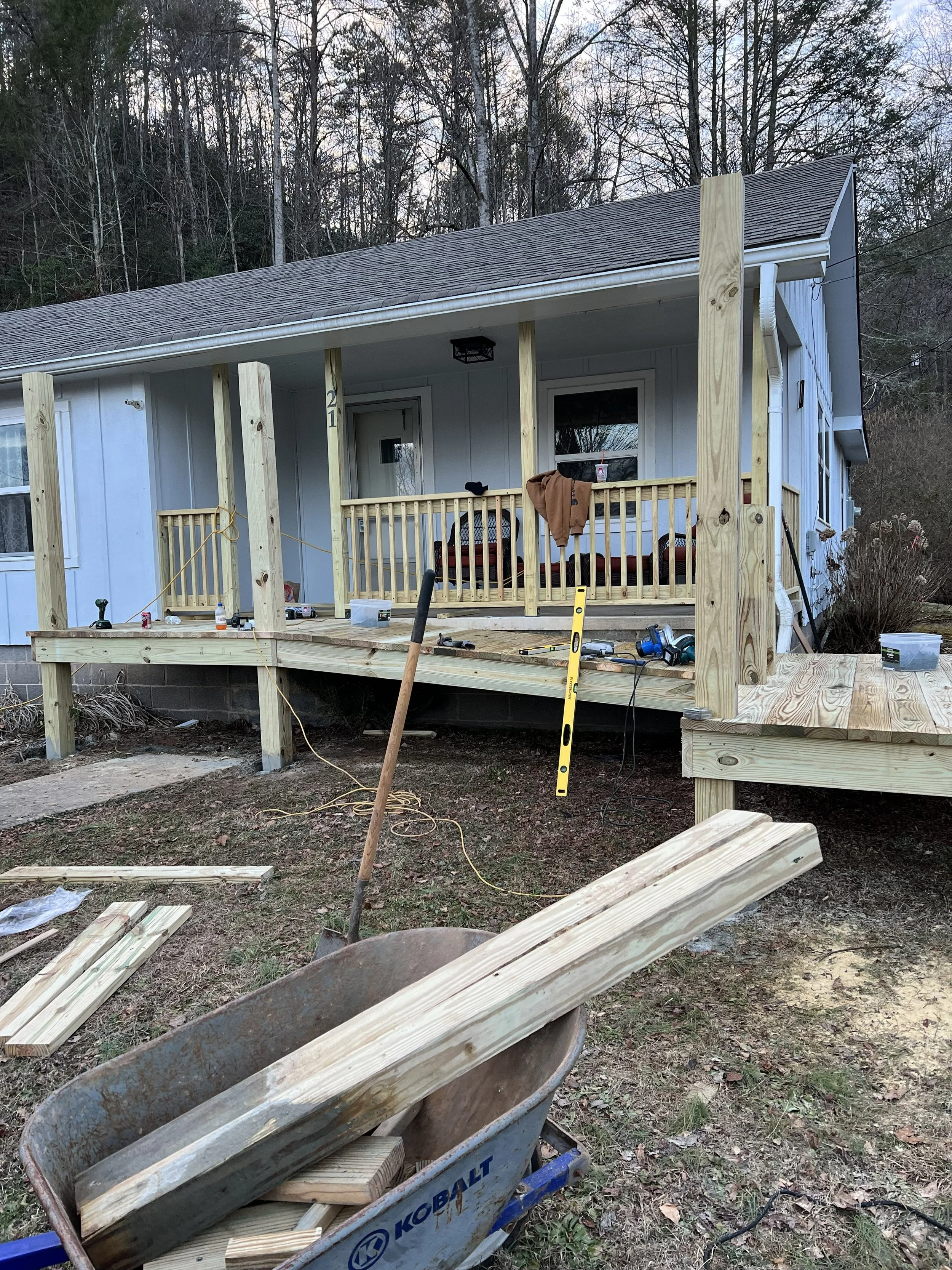 Construction site with a small wooden porch attached to a house, showing an unfinished railing and support posts, with tools and materials scattered around, and a wheelbarrow filled with wood pieces in the foreground, against a background of trees an