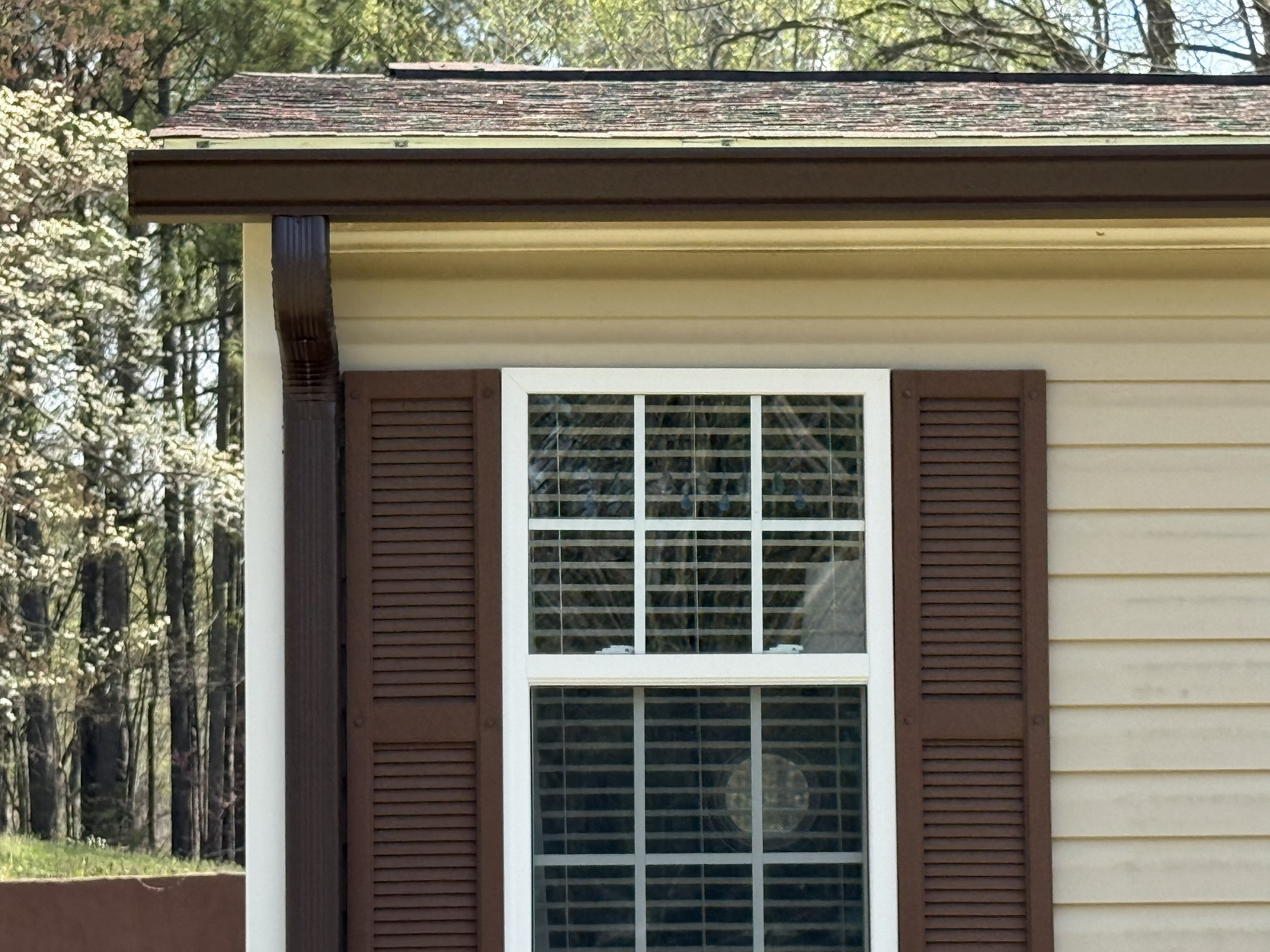The photo shows a house with beige siding, a window with brown shutters, a window air conditioning unit, a roof with shingles, and trees in the background.