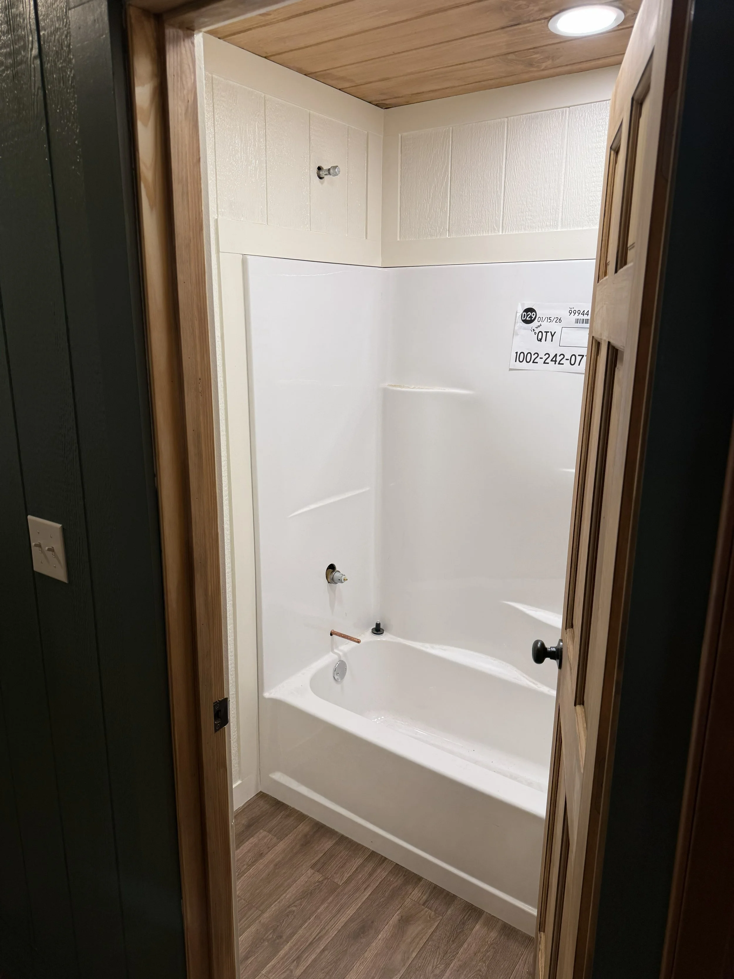 Empty bathtub inside a shower stall with wood-paneled ceiling and door frame, tile walls, and a floor with wood-look planks.