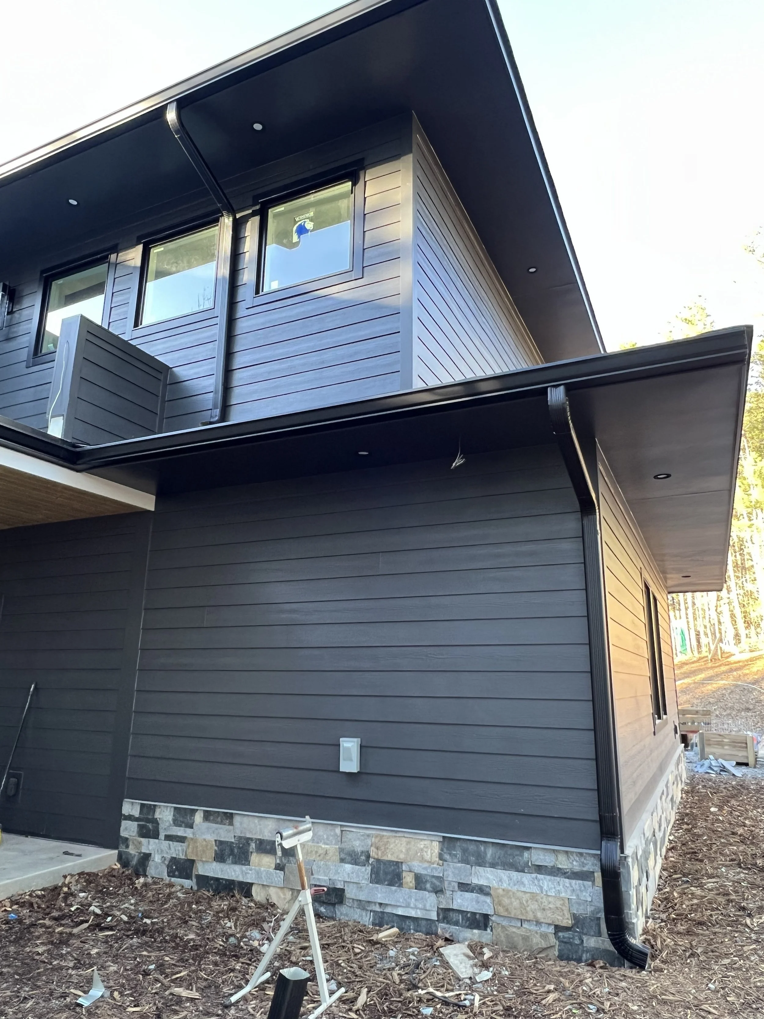 Side view of a modern house with black siding, a stone foundation, and a black gutter system. Construction tools and debris are visible around the base of the house.