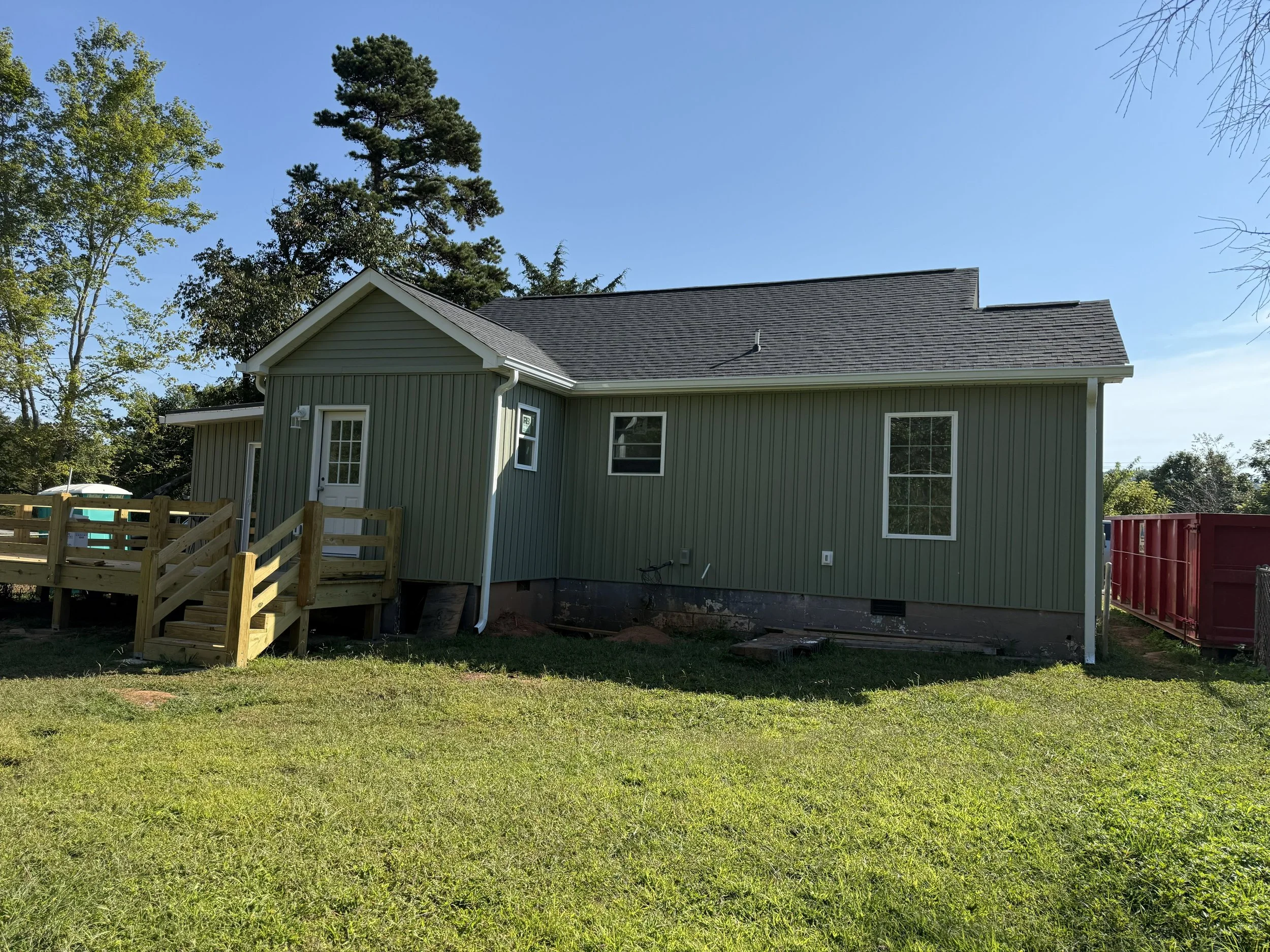 A house with green siding and a gray roof, surrounded by trees and a wooden deck at the back, under a clear blue sky.