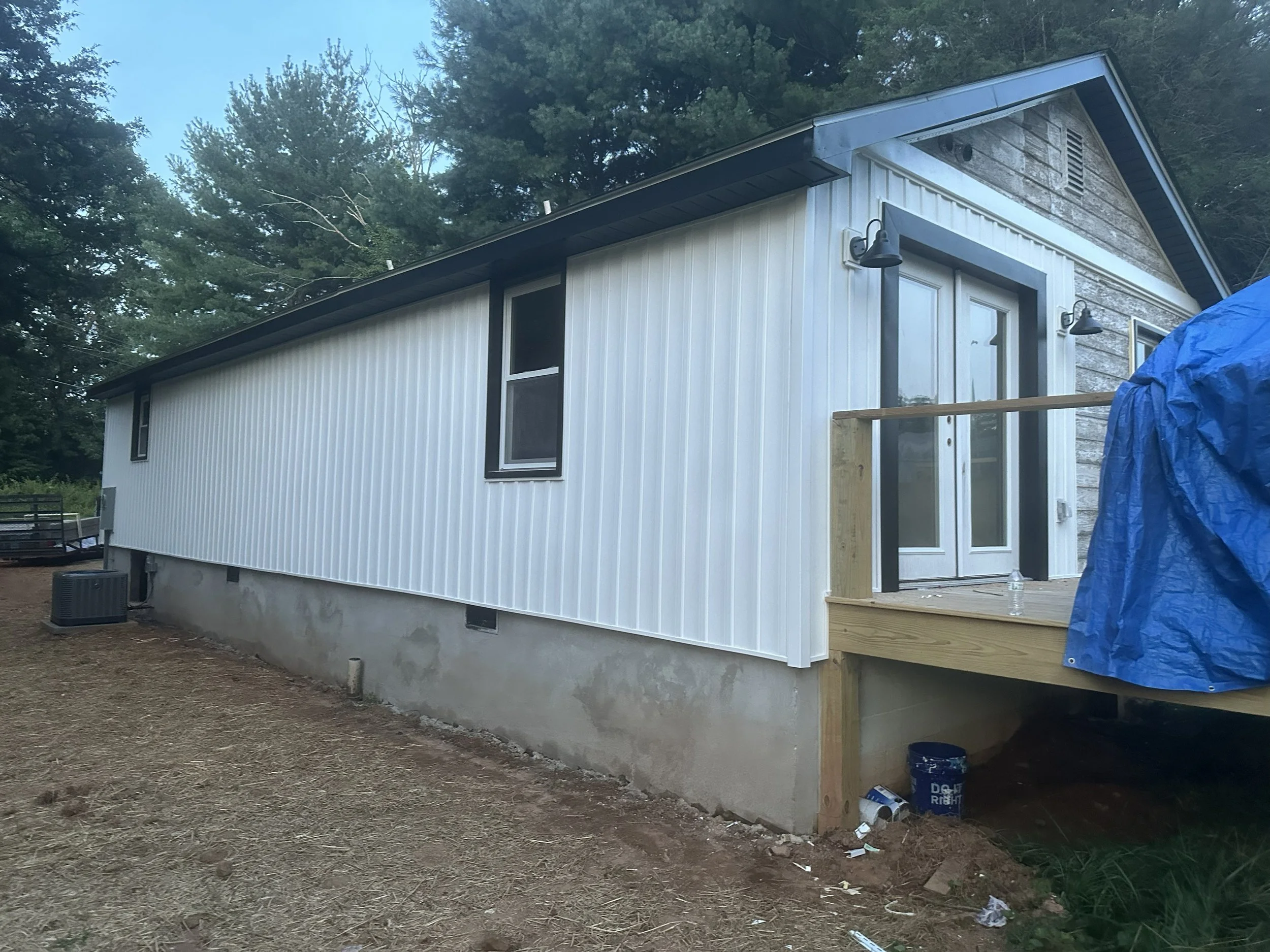 A house under construction with white siding, large windows, and outdoor wall-mounted lights. The house has a deck with a wooden railing, and the surroundings include trees and construction materials and debris on the ground.