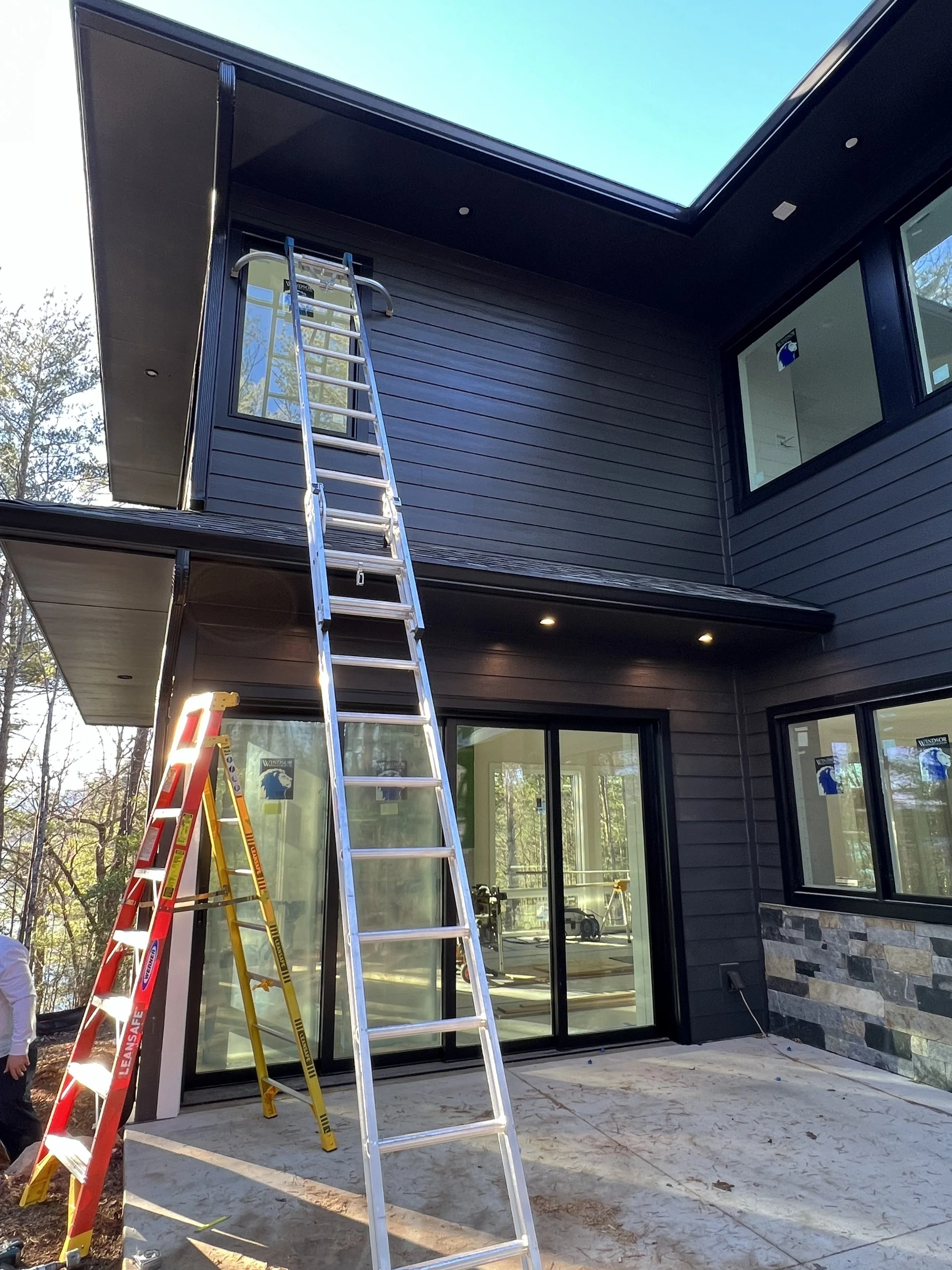 Ladders leaning against the exterior of a modern black house with large windows, an upper outdoor balcony, and construction or renovation work in progress.