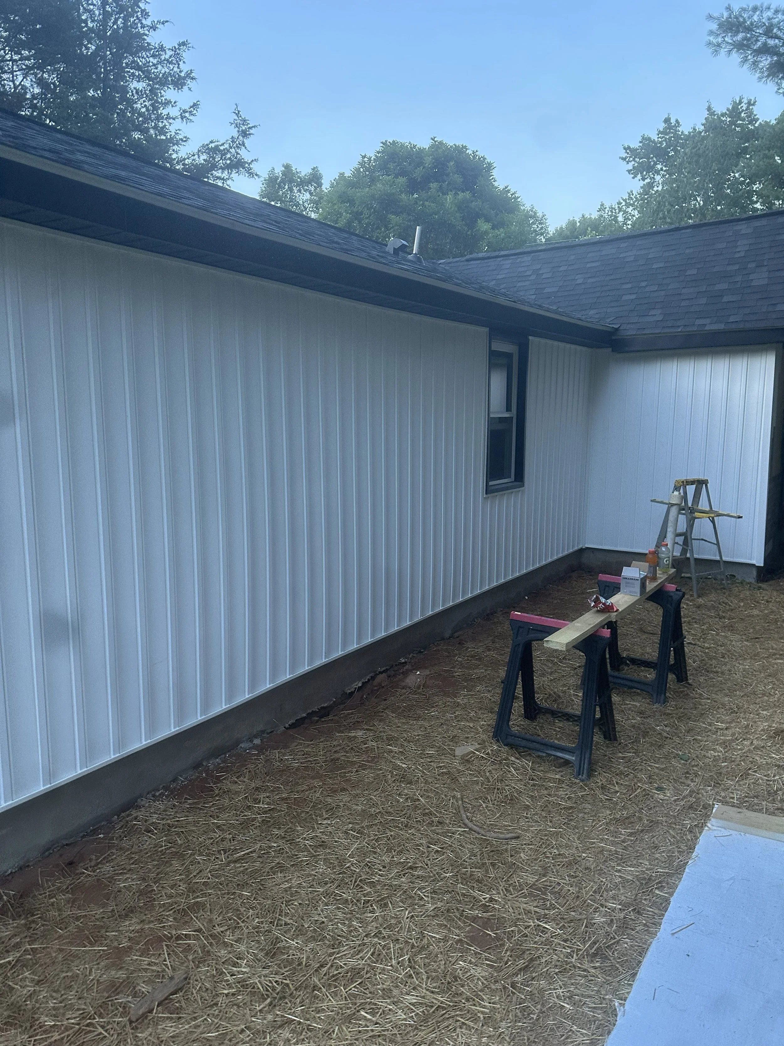 Exterior of a house with white metal siding, a black roof, and a small window. There is a ladder, tools, and construction materials on the ground indicating ongoing construction or renovation. The ground is covered with straw or hay.
