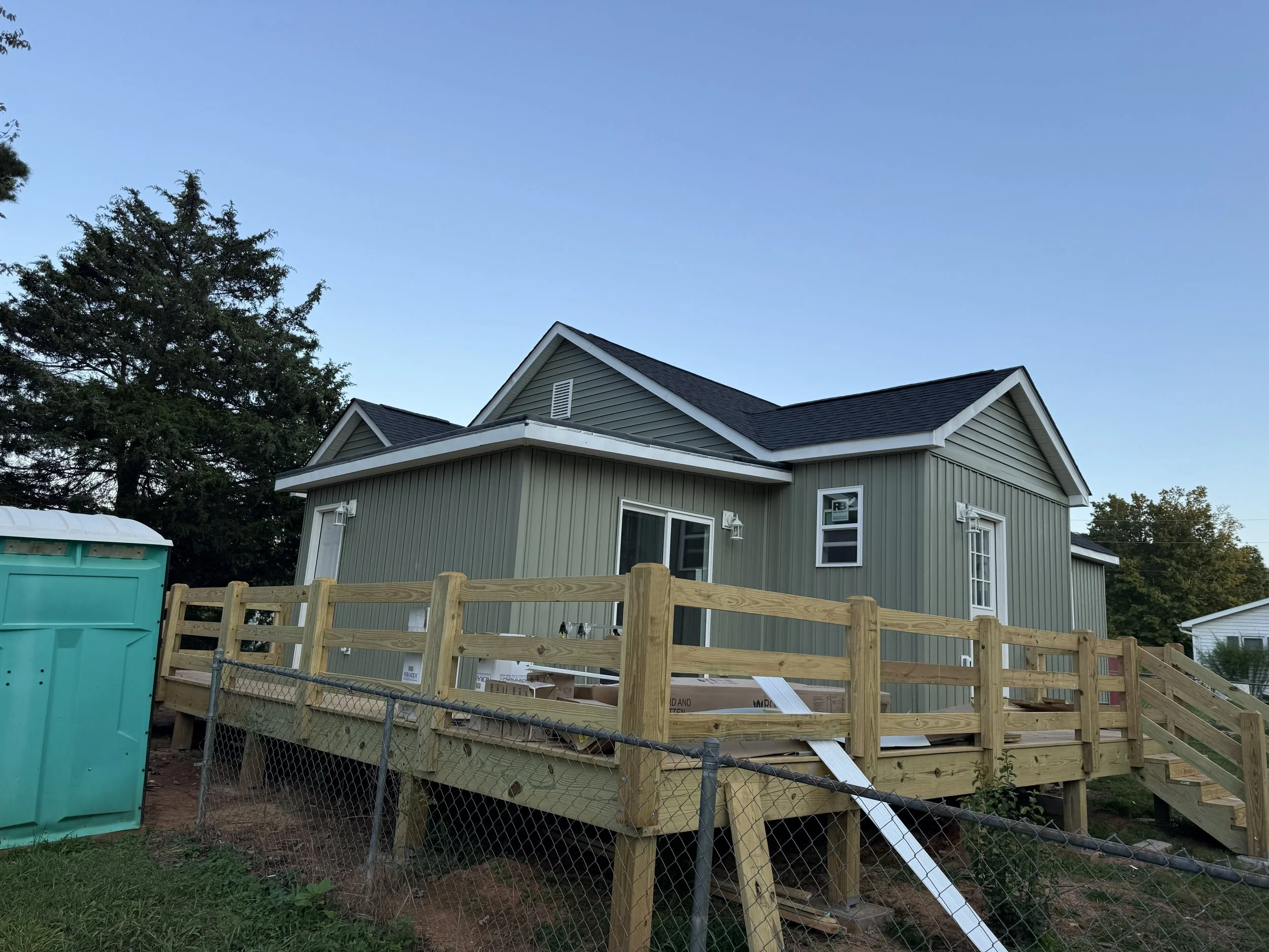 A house under construction with a wooden deck, green siding, multiple windows, and a dark roof. There is a large tree adjacent to the house, a green portable toilet, and a chain-link fence surrounding the area.